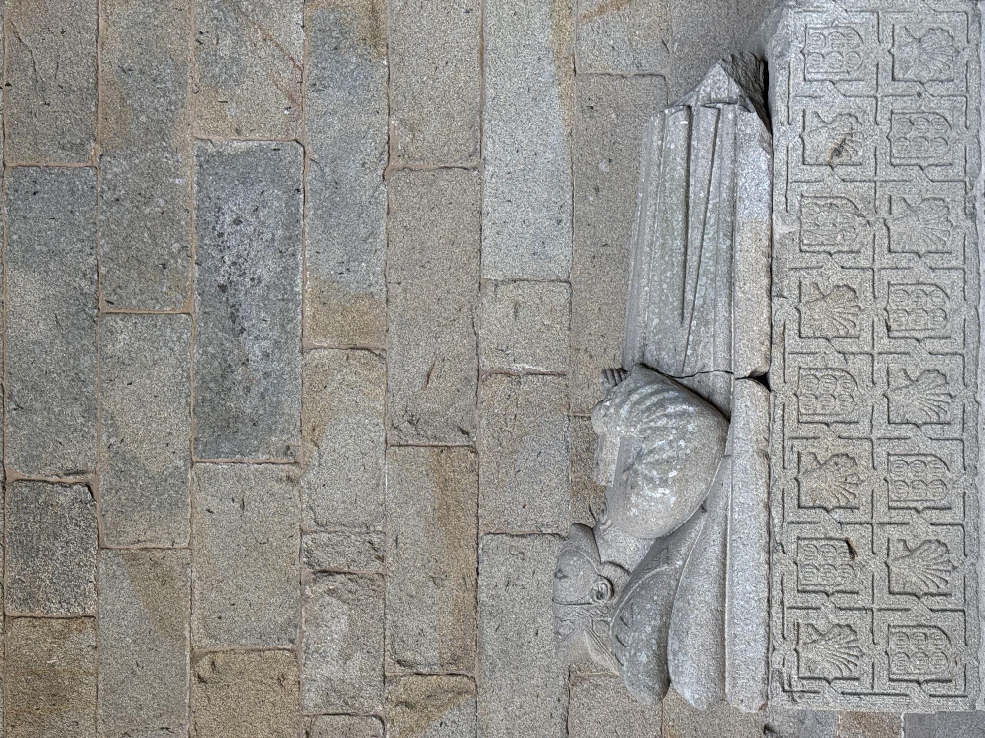 The image shows a stone effigy of a reclining figure, possibly a knight or noble, situated on a carved stone sarcophagus. The figure is lying on its back, with its head resting on a pillow. It is adorned with what appears to be a crown or elaborate headdress, and armor or ornate clothing. The sarcophagus itself is decorated with a repeating pattern of shields and other heraldic or decorative motifs carved into its surface. Behind the sarcophagus is a rough-hewn stone wall, characteristic of ancient buildings. The lighting suggests an outdoor or semi-enclosed space, possibly a cloister or courtyard. This effigy is located in the Cathedral of Santiago de Compostela, Spain, specifically within the cloister.