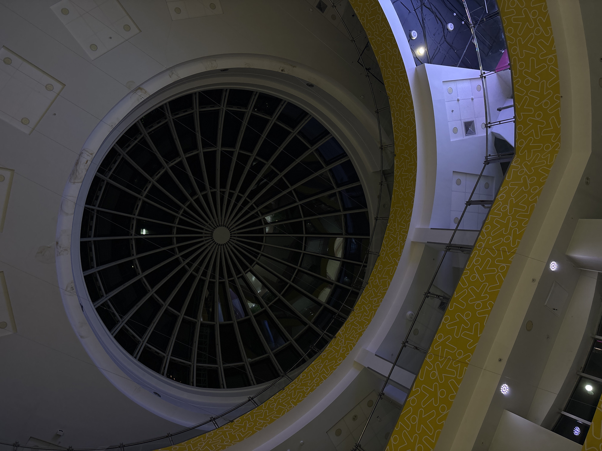 The image depicts the interior of the "iMall Esplanada" shopping center in Perm, Russia, focusing on the large circular skylight and the adjacent architectural elements. The skylight is composed of a grid-like pattern, providing a view upwards. Around the skylight and along the walls are sections of yellow decoration with an abstract stick-figure design. The walls are predominantly white, with some sections illuminated by spotlights. Scaffolding and support structures are visible in the background. The scene is captured from a low angle, looking upwards towards the ceiling and skylight.
