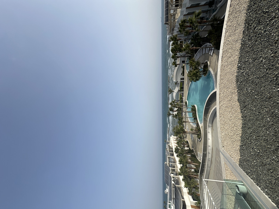 This image captures an elevated, deserted coastal resort scene in Oroklini, Cyprus, under a clear, cloudless sky. The immediate foreground consists of a dark grey gravel surface, transitioning to a lighter beige gravel pathway, bordered by a low white-capped architectural element. A glass railing and a grey cylindrical planter containing greenery are visible on the far left foreground.
The midground showcases a large, irregularly shaped swimming pool filled with light blue water, surrounded by extensive paved areas and numerous green palm trees. Adjacent to the pool are several modern, multi-story white buildings with distinctive curved roofs, many featuring integrated solar panels. Unoccupied lounge chairs are scattered around the pool area and pathways.
The background displays the Mediterranean Sea, characterized by blue-green water with visible waves breaking near the shoreline. A faint outline of a distant vessel is discernible on the sea surface towards the right. No individuals, actions, or interactions are present in the frame. The environment is illuminated by bright daylight.