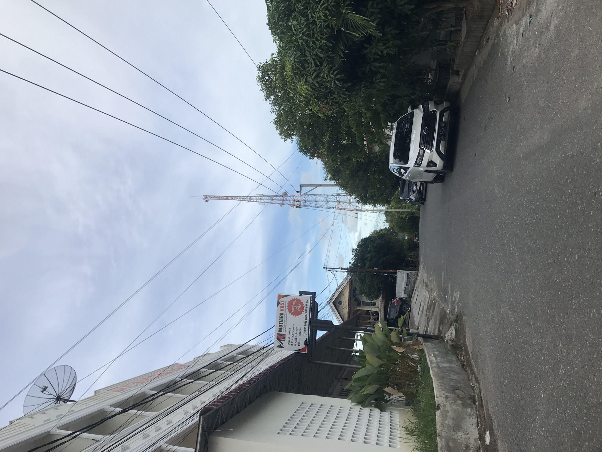 The image depicts a street scene in Lhokseumawe, Indonesia. A white Toyota Innova car with license plate BL 1432 NU is parked on the right side of the asphalt road. In the background, a lattice-like communication tower rises against a cloudy sky. Overhead electrical wires crisscross the sky. A building on the left features a satellite dish. A sign for "Mutiara Kost" is visible on the building, advertising daily, weekly, monthly, and annual room rentals. A second car is parked further down the street. The environment consists of buildings, trees, and foliage, indicating a residential or commercial area.