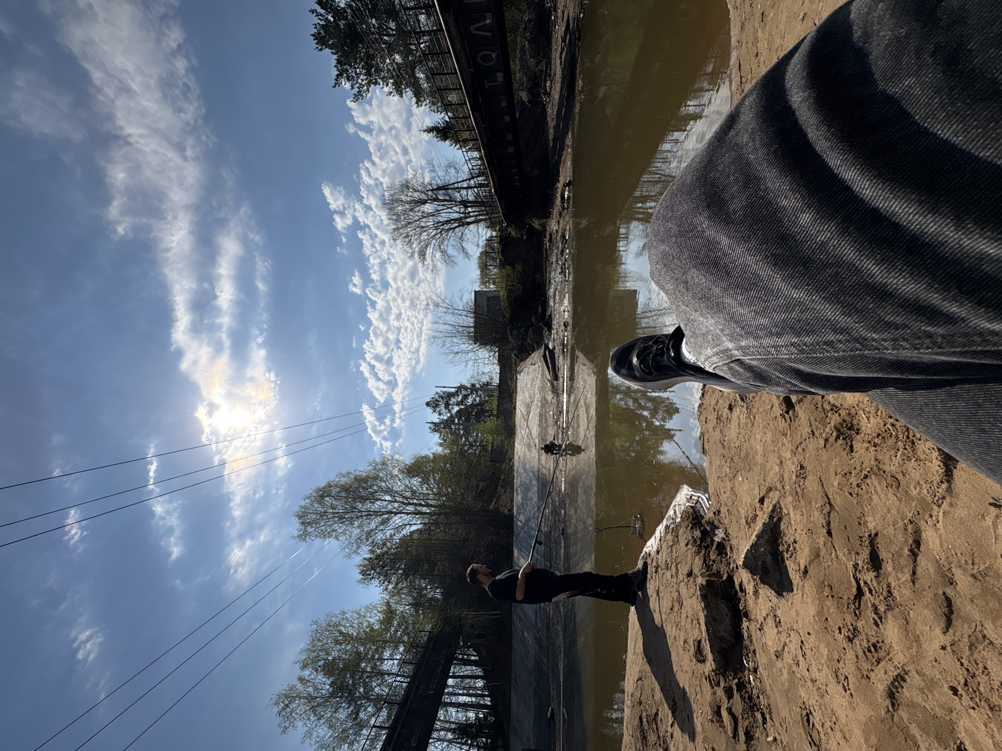 The image shows a person standing on a muddy embankment beside a canal in Chaikovsky, Russia. The canal is filled with murky water reflecting the sky and surrounding trees. In the background, there is another person fishing, using what appears to be a long pole, along with a fishing net and other fishing equipment. Overhead, power lines stretch across the sky. The sky is blue with streaky clouds and bright sunlight. The person in the foreground wears dark clothing and denim pants. A concrete structure with the word "LQAR" printed on it is visible on the far side of the canal. Trees line the canal banks, with some buildings visible in the distance. The embankment appears to be made of sand and mud.