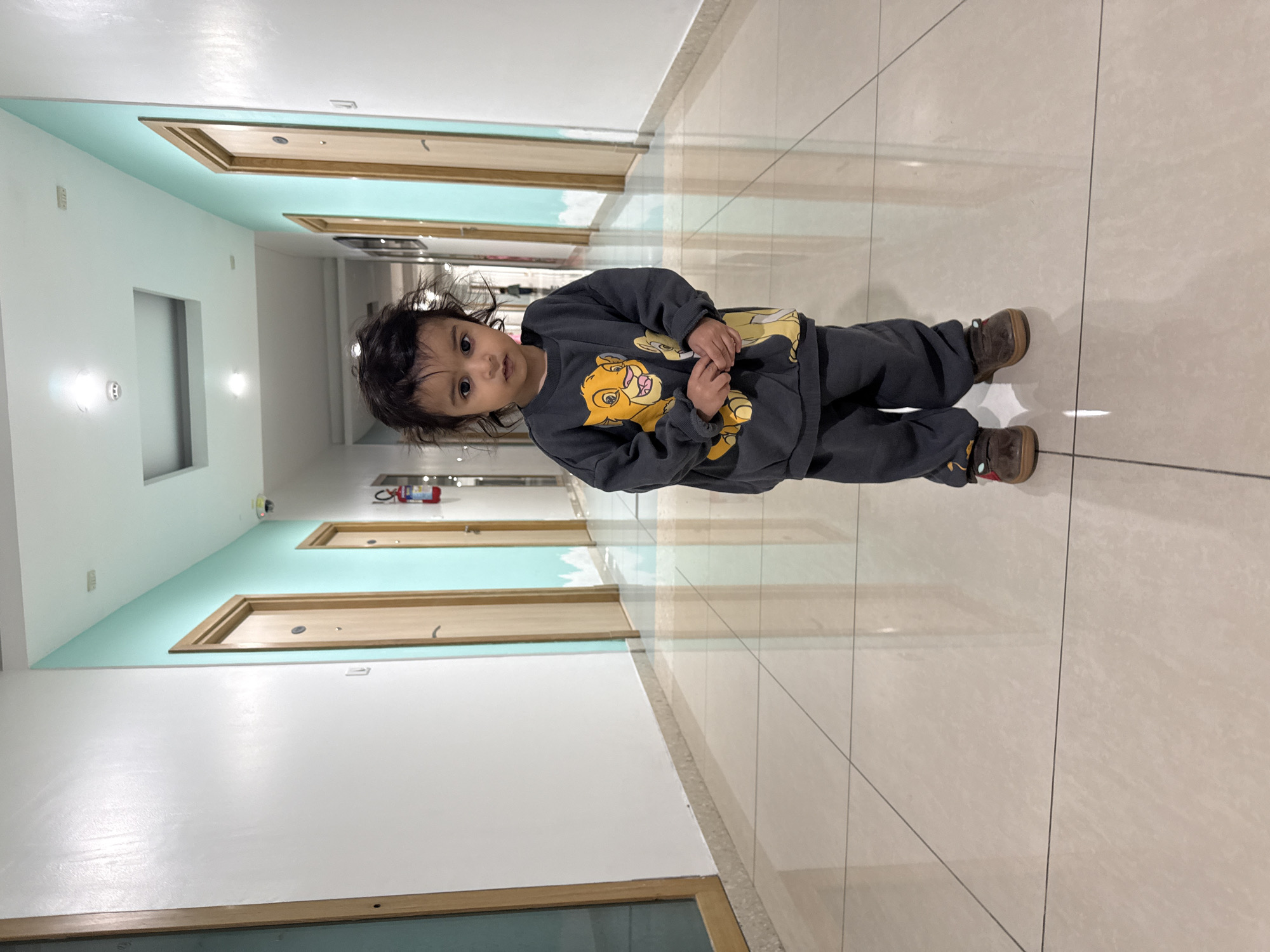 A young child wearing a gray Lion King-themed sweatshirt and brown shoes stands in a hallway. The hallway has white walls with a light teal accent, and wooden-framed doors. The floor is tiled in a light beige color. The location is likely an interior space in Marunji, India.