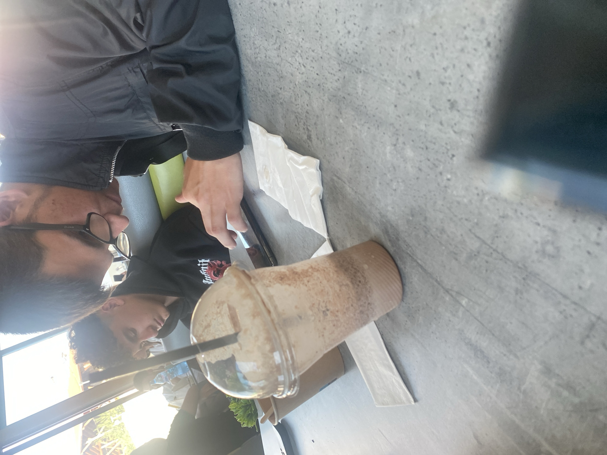 The image shows a table with a clear plastic cup containing a blended beverage, likely a milkshake or smoothie, with a black straw. To the left of the cup is a small potted plant. Behind the cup, a person wearing a black hoodie is seated, looking down. Another person wearing a black jacket is to the right, also looking down, with glasses on. A white napkin is placed on the table between the cup and the person in the black jacket. The scene appears to be indoors, possibly a cafe or restaurant, with natural light entering from windows or an open space. The table surface is a grey, textured material. The image does not contain any information that would suggest the location is in Ablanitsa, Bulgaria.