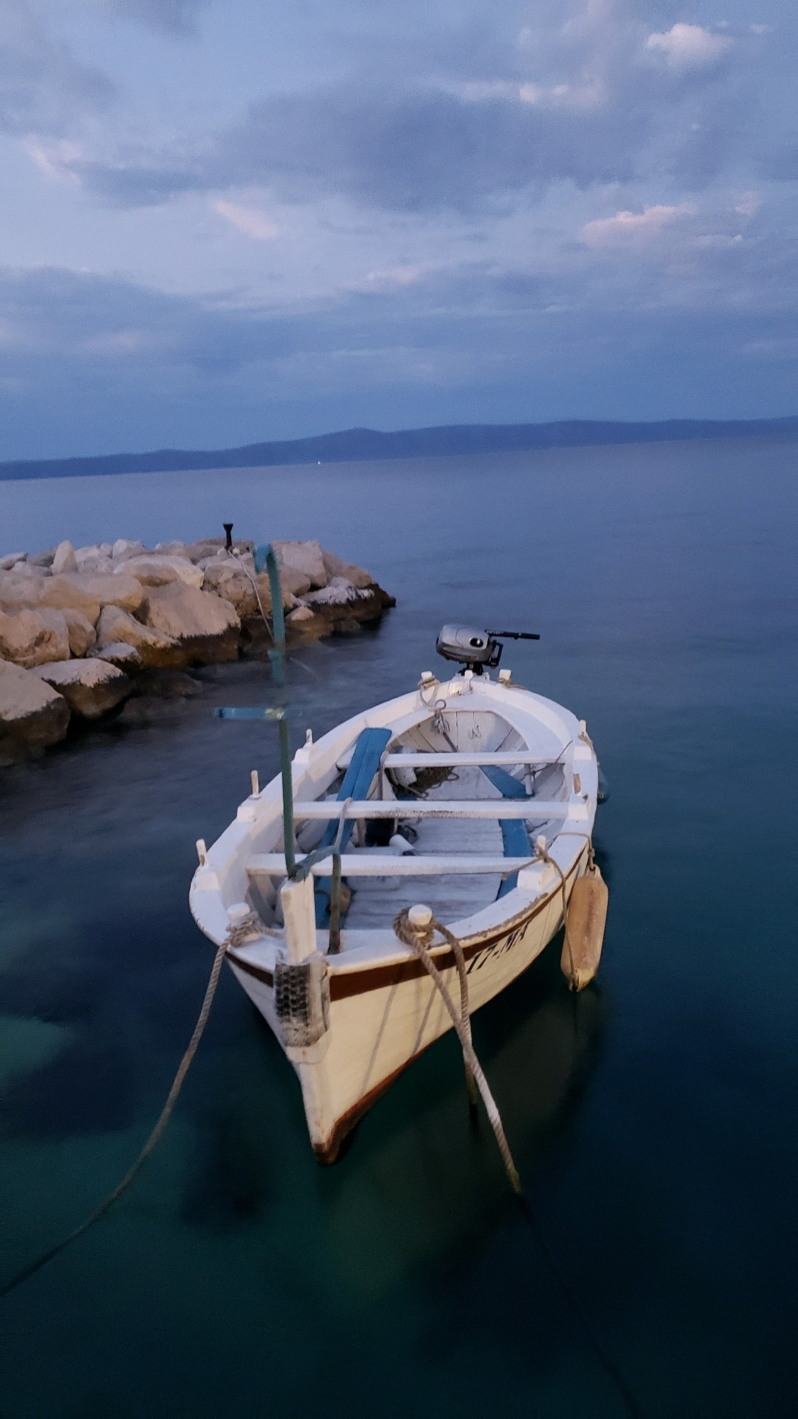 This is a vertical, high-angle shot of a white fishing boat docked in the clear blue waters of Općina Tučepi, Croatia. The boat is empty and tethered to a small, rocky pier. The sky above is overcast with shades of blue and purple, suggesting it is either dusk or dawn. The sea is calm, with the underwater structure of the pier visible through the translucent water. A small outboard motor is mounted on the stern of the boat, and blue seats are visible inside. A fender hangs from the side of the boat, and ropes are used to secure it. The image is serene, with no people present.