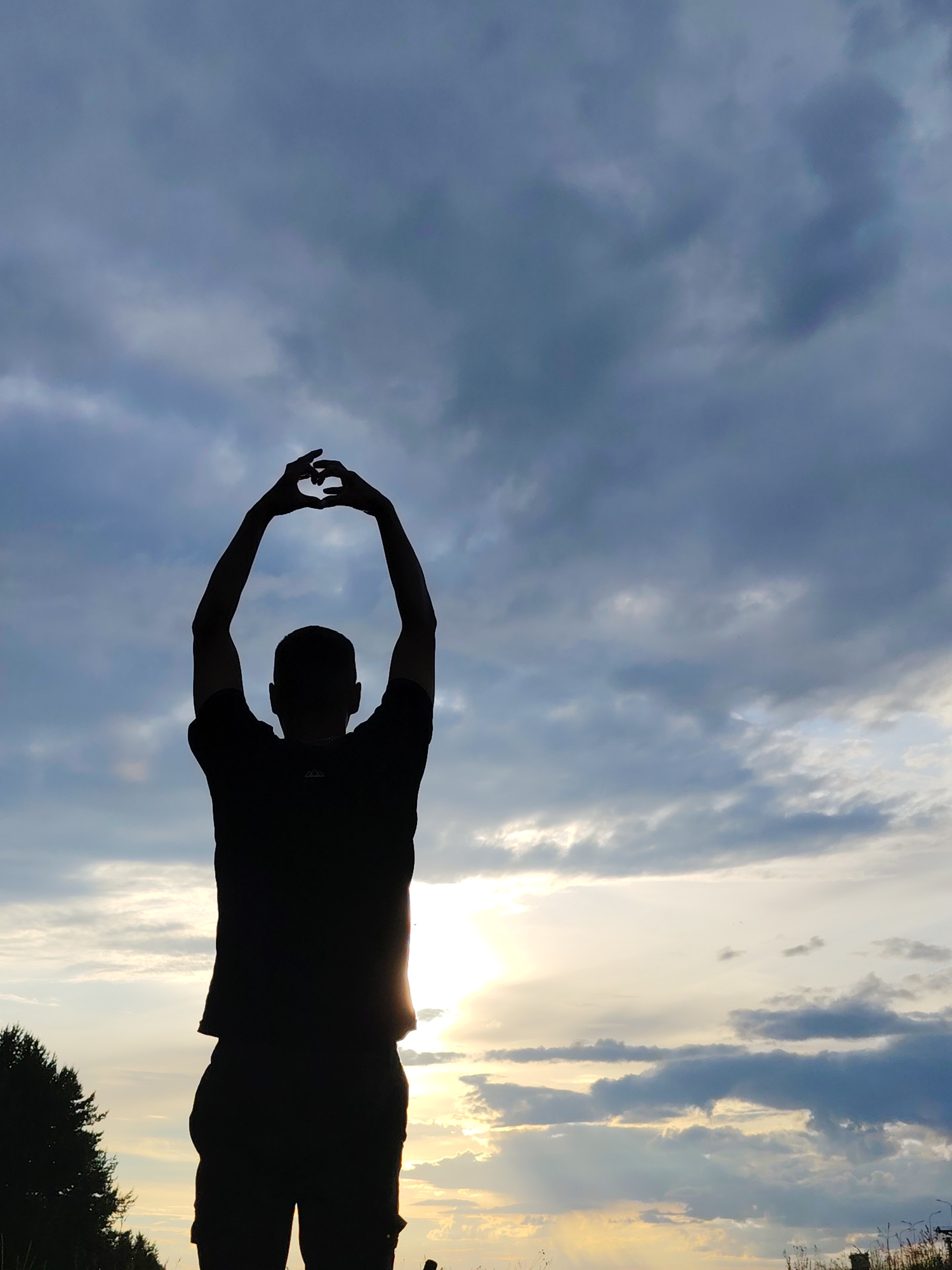 The image features the silhouette of a person with their arms raised above their head, forming a heart shape with their hands. The subject is viewed from behind, wearing a t-shirt and pants. The setting is outdoors, with a dramatic sky filled with clouds, suggesting either dawn or dusk. The sun is partially visible, casting a warm glow and creating a lens flare. To the left of the frame, a line of trees is visible, hinting at a natural or rural landscape. The overall mood is peaceful and contemplative. No text is visible in the image.