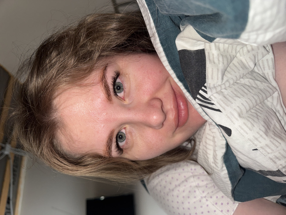 The image depicts a female subject in an indoor setting, identified as the city of Gornyi (Dvurechenskoe S/P), Russia. The subject, a woman with light brown, slightly wavy hair and light blue-grey eyes, is positioned facing the camera, seemingly lying down in a bed. Her face is in the foreground, displaying a slight smile with her lips slightly parted. Her skin appears fair with some redness on the cheeks and forehead. She has prominent eyelashes, consistent with enhancements such as extensions, and well-defined eyebrows. She is partially covered by a duvet or blanket featuring a patterned cover in white, light grey, dark teal, and black geometric or abstract designs. A white pillow with faint purple polka dots is visible on the left side of the frame, supporting her head. The background is indistinct and dark, with a blurred dark rectangular object (possibly a television or monitor) visible in the upper left and faint structural elements, potentially ceiling joists or pipes, in the upper center. The scene is lit, illuminating the subject's face directly.