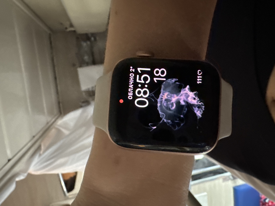 The image displays a human left forearm and wrist, with light skin tone, wearing a rose gold-toned Apple Watch with a light-colored silicone band. The watch face is active, showing "ОБЛАЧНО 2°" (Cloudy 2°C) at the top, the time "08:51," the number "18," and a vibrant, translucent purple and pink jellyfish animation in the center. A "111♡" icon is present at the bottom right of the screen. The background is an out-of-focus indoor environment, featuring light-colored overhead panels or structures, and a white material draped over an unseen object on the left, possibly within a vehicle or room in the city of Gryazi, Russia.