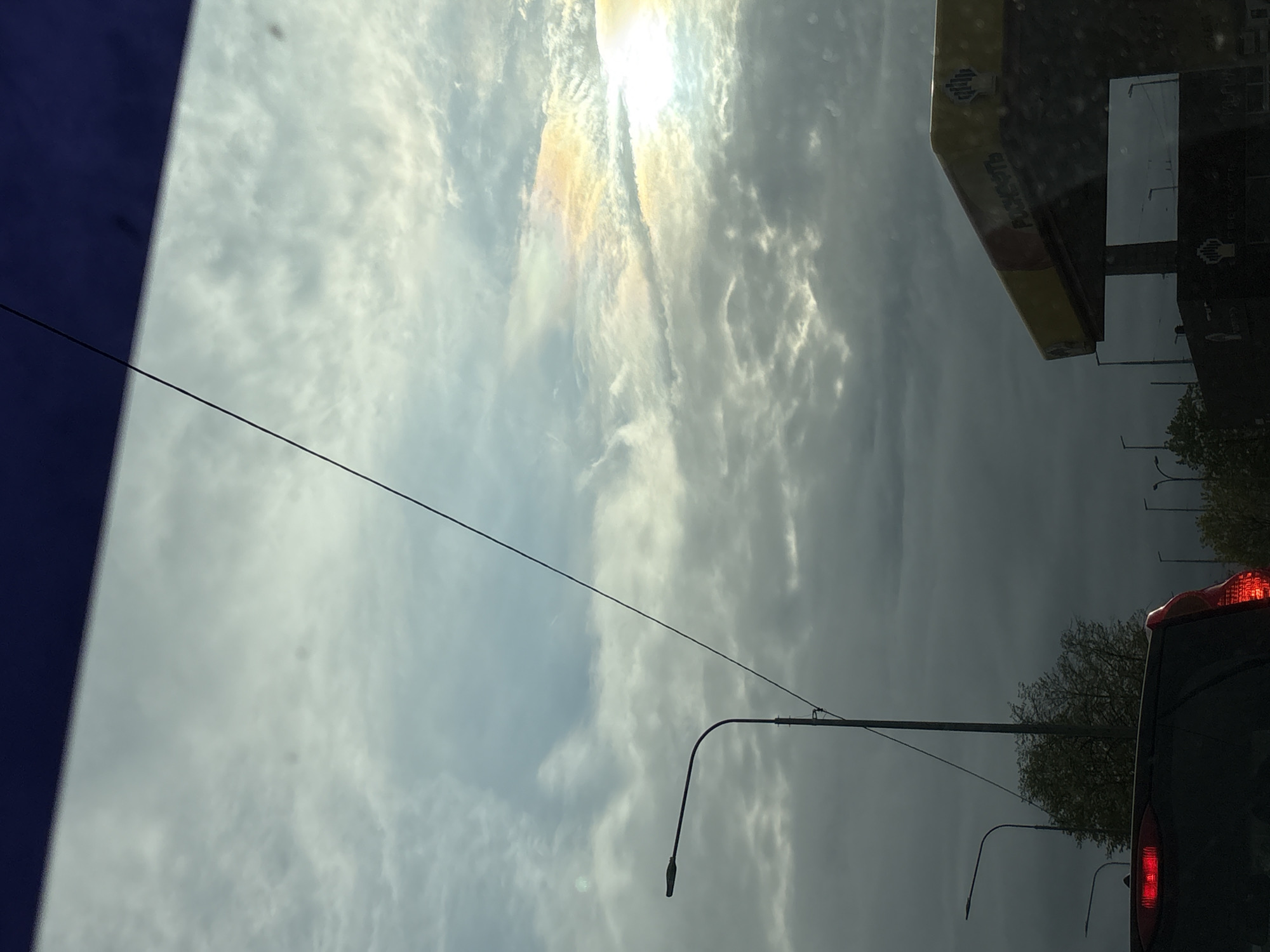 The image depicts a view from inside a vehicle, possibly in Saint Petersburg, Russia, given the Cyrillic lettering on the gas station sign visible through the windshield. The sky is overcast with a bright area where the sun is obscured by clouds, creating a rainbow-like effect. A power line stretches across the sky. A street lamp is visible. The rear taillights of a car are lit, indicating the vehicle is potentially braking or stopped. Trees are in the background. The color of a part on the left of the photo indicates that the picture has been taken from within a vehicle.