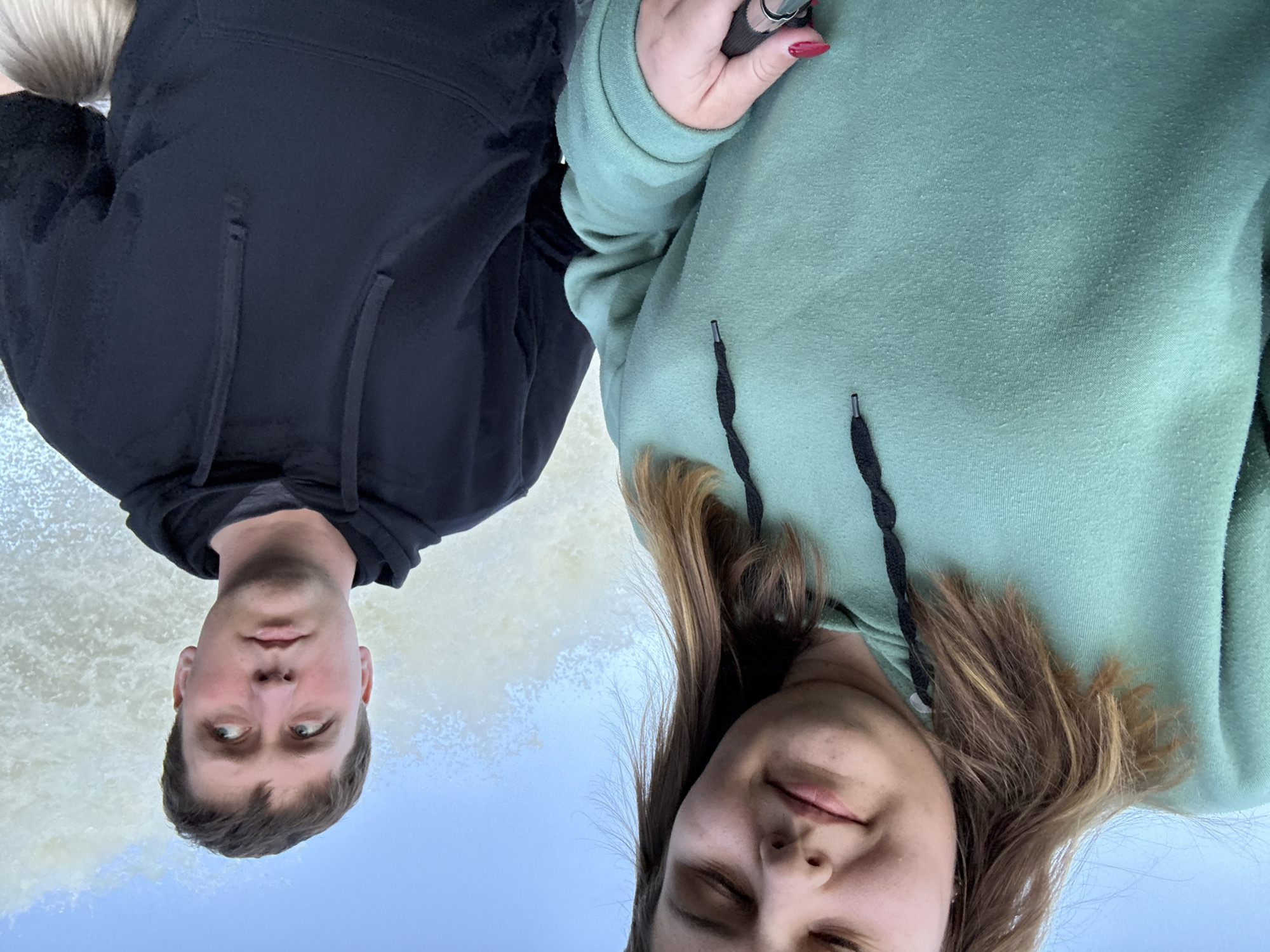 Two individuals are positioned in front of a landscape that includes a body of water and sky. The person on the left, appearing male, wears a black hooded sweatshirt. The person on the right, appearing female, wears a light green hooded sweatshirt. The individuals are situated with their faces close together. The location is Chernushka, Russia.