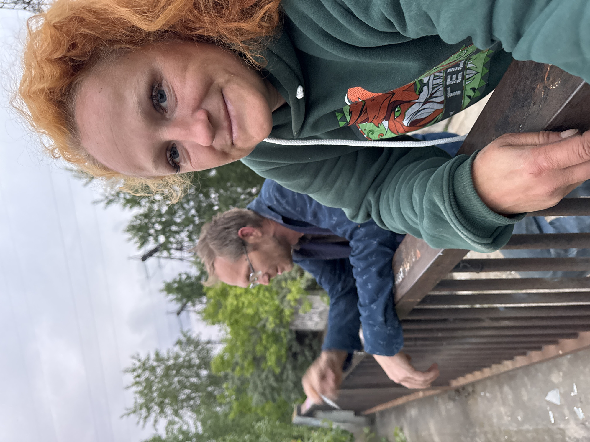 The image shows a woman with red, curly hair and a man with glasses. The woman is in the foreground, facing the camera with a smile. She is wearing a green hooded sweatshirt with a graphic of a fox on it. The man is in the background, leaning on a railing, and appears to be smoking a cigarette. They are standing on a bridge or walkway with a metal railing. In the background, there are trees and a cloudy sky. The location is identified as Chaikovsky, Russia.
