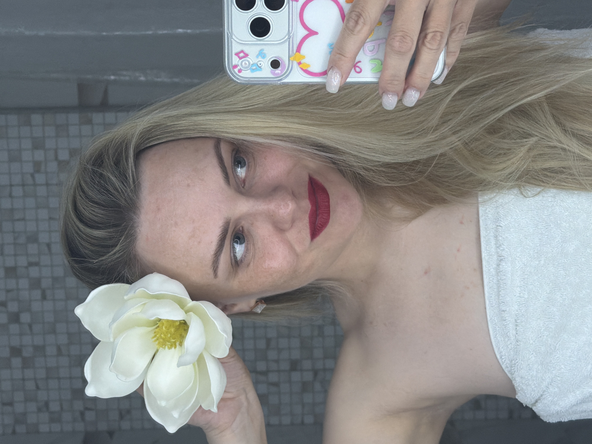 A light-skinned woman with blonde hair is shown in a selfie format, holding a white flower near her face. She has red lipstick and is wearing a white towel. Her phone, encased in a clear case with a colorful design, is visible. The background appears to be a tiled wall. It is not possible to identify the location as Votkinsk, Russia with the available information.