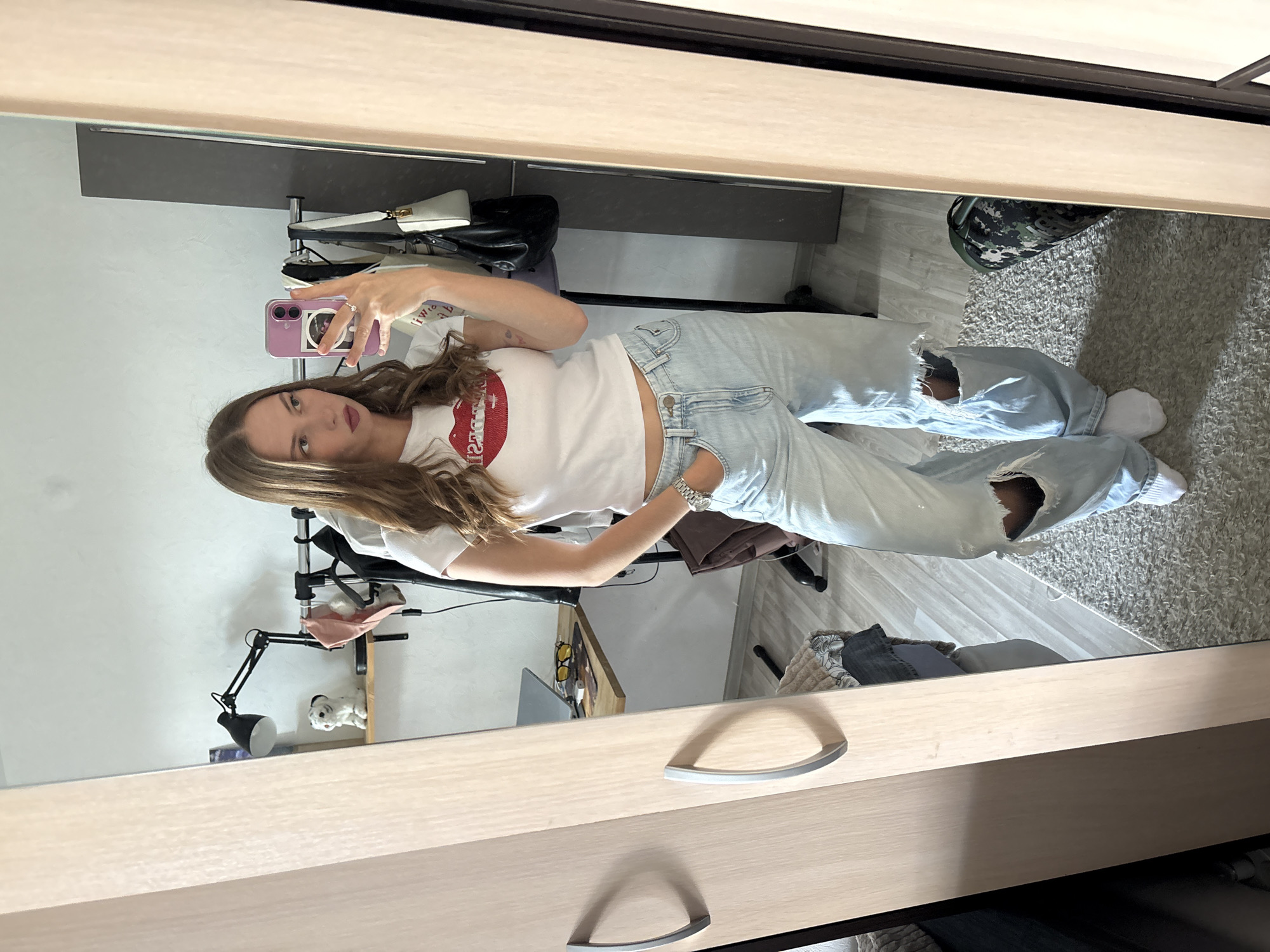 A young woman with long brown hair is taking a mirror selfie in what appears to be a bedroom or dressing area. She is wearing a white t-shirt with a red lip graphic and light blue, distressed jeans. She is holding a purple smartphone in her right hand, angled to capture her reflection.
The background, seen in the mirror's reflection, shows a closet with clothes hanging and bags on a shelf. To the right, a gray carpet covers the floor, and a camouflage-patterned shoe is visible. A desk with a lamp and what looks like a laptop is also reflected. There is a small white stuffed dog with black ears on a shelf.
The room appears to be indoors, with artificial lighting from a lamp and overhead fixtures. The overall impression is casual and personal, possibly a moment captured while getting ready or simply documenting an outfit. The woman's expression is neutral, and the image seems to have been taken during the daytime, judging by the light. There is no visible text in the image.