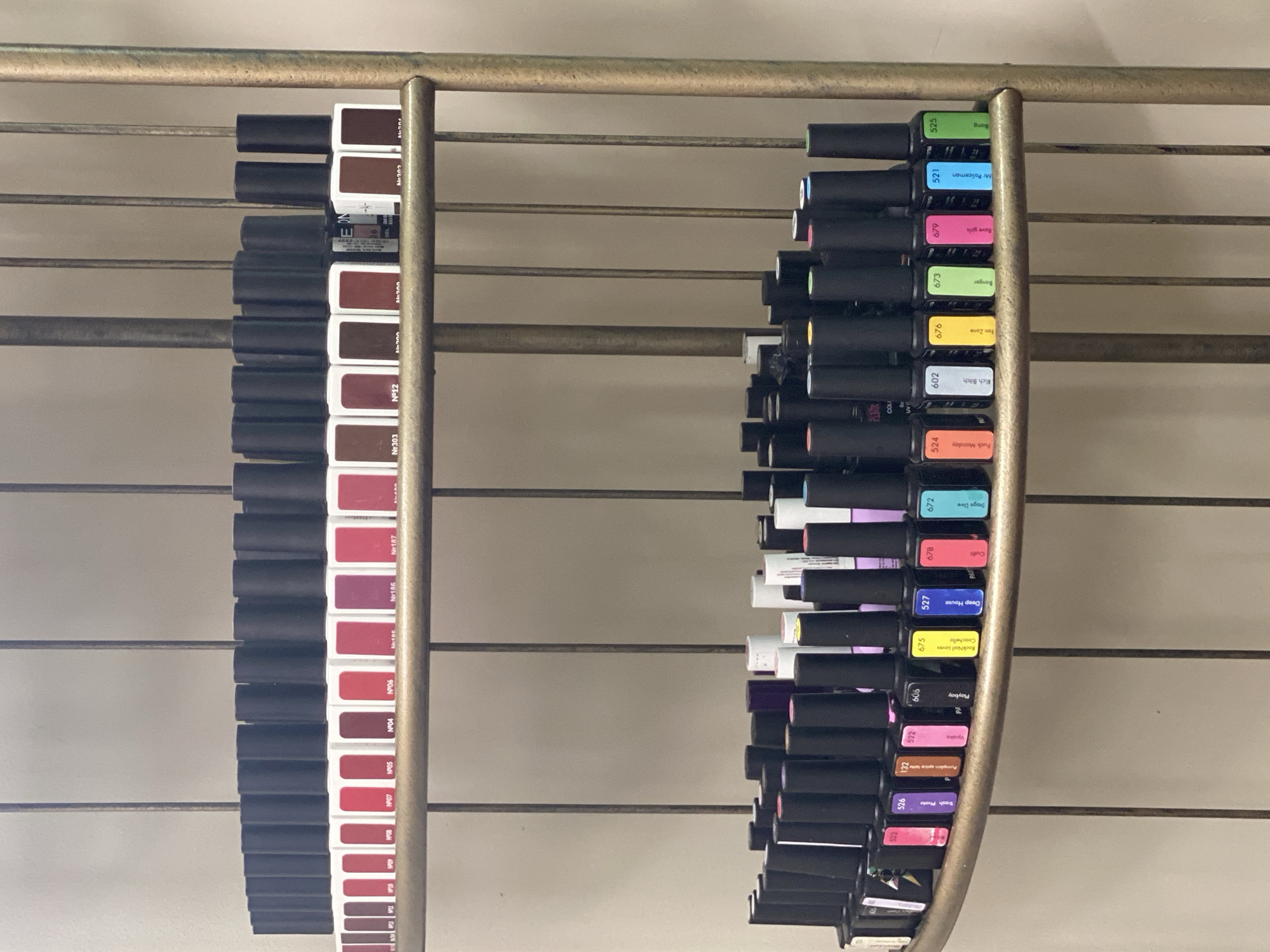 The image shows two metallic racks filled with nail polish bottles. The rack on the left displays bottles with brown and red color samples on their labels, while the rack on the right features bottles with a variety of colors. Each bottle has a black cap and a label indicating the color name and code. The racks are mounted on a wall with horizontal slats. The nail polish color names visible on the rack to the right include: "Trash Photo," "Pumpkin Spice Latte," "Vpiske," "Playboy," "RockNail Lovers Coachella," "Deep House," "Outfit," "Stage Dive," "Fuck Monday," "Rich Bitch," "Fan Zone," "Banger," "Rave girls," "Mr Policeman," and "Bong". The location in Sochi, Russia is not discernable from the image.