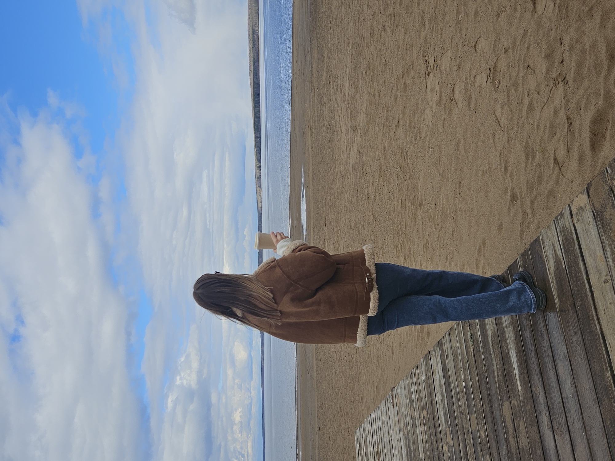 A person stands on a wooden boardwalk overlooking a beach and a body of water, likely a lake or reservoir. The boardwalk is made of light-colored wooden planks running vertically in the image. The beach consists of sand that transitions from darker brown near the water to a lighter shade closer to the boardwalk. The water appears calm and reflective, with a strip of land visible in the distance on the opposite shore. The sky is blue with scattered white clouds. The person is dressed in jeans and a brown jacket with a light-colored lining, holding a small object in their hand, possibly a cup or container. The location is the city of Chaikovsky, Russia.