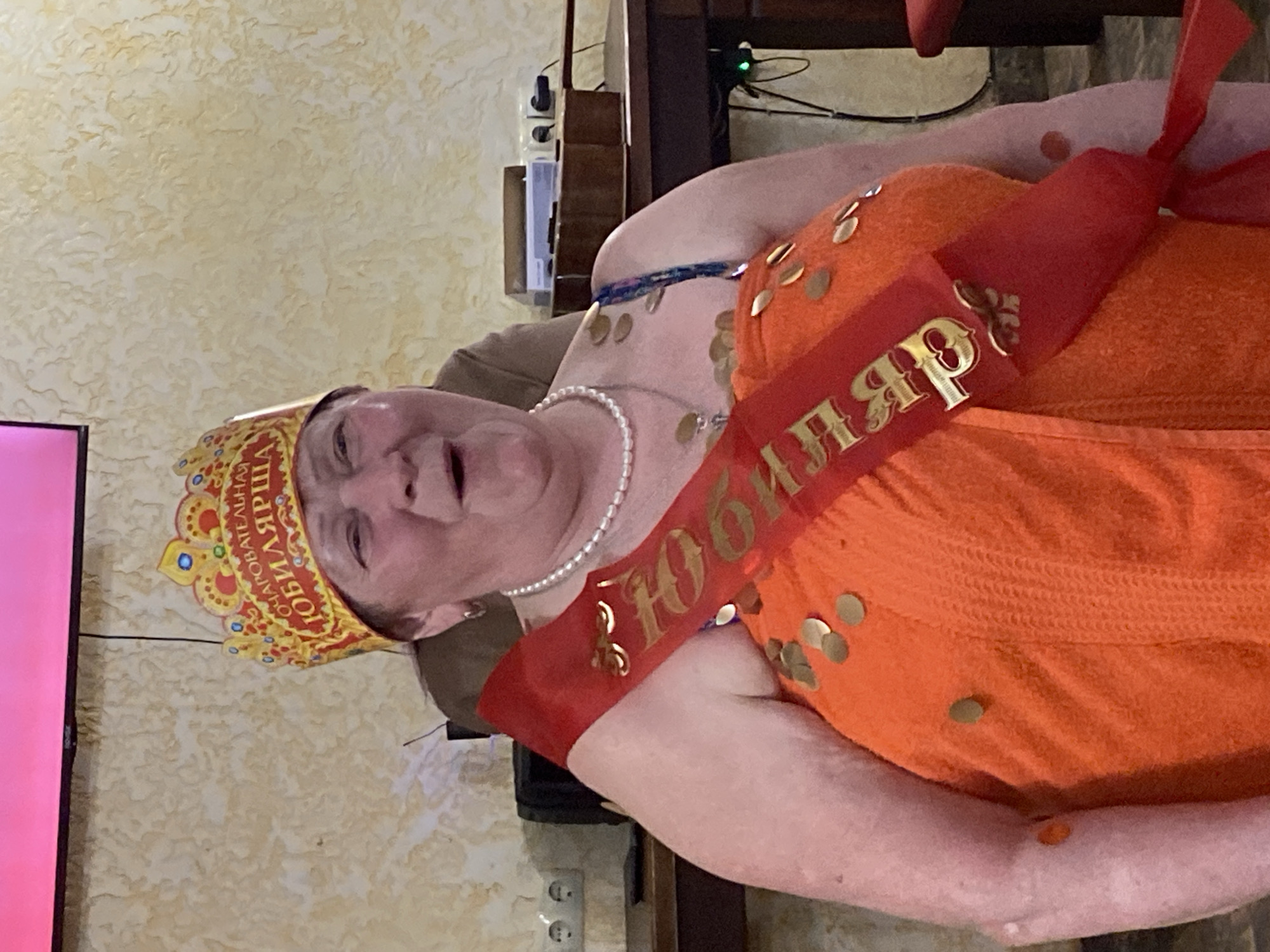 An older Caucasian woman is photographed indoors, likely in a residence. She is wearing a crown that reads "ОЧАРОВАТЕЛЬНАЯ ЮБИЛЯРША" (Charming Anniversary Girl) and a red sash that says "ЮБИЛЯРША" (Anniversary Girl) in Cyrillic lettering. The sash is decorated with metallic gold circles that match the ones on the orange dress or wrap the woman is wearing. She also has a pearl necklace. The woman's position suggests she is in Chaikovsky, Russia. In the background, there is a wall with a textured, beige pattern. There is also a dark wood cabinet and an outlet.