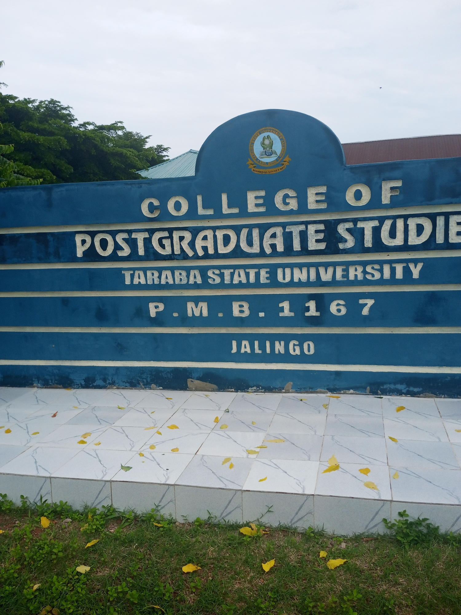 The image shows a blue sign with white lettering that reads "COLLEGE OF POSTGRADUATE STUDIES TARABA STATE UNIVERSITY P.M.B. 1167 JALINGO". The sign is mounted on a blue wall, and in front of the wall is a tiled area with fallen yellow leaves. Behind the sign, a building with a green roof and red trim is visible, along with some trees and a cloudy sky. The sign is located in Jalingo, Nigeria.