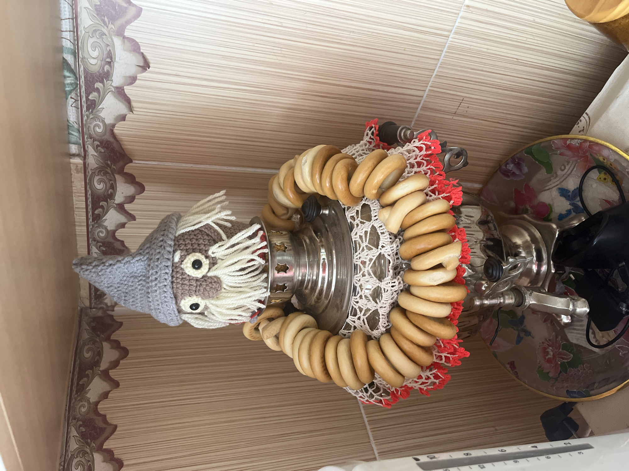 A knitted doll with a grey pointed hat, white hair, and a brown face is placed on top of a metal samovar. The doll is adorned with a garland of ring-shaped bread rolls. The samovar is decorated with a white crocheted doily and a red and white fringe. The entire setup sits on a decorative tray with floral patterns. The background consists of beige tiled walls and a light-colored shelf. The scene is located in the city of Сухринское, Russia.