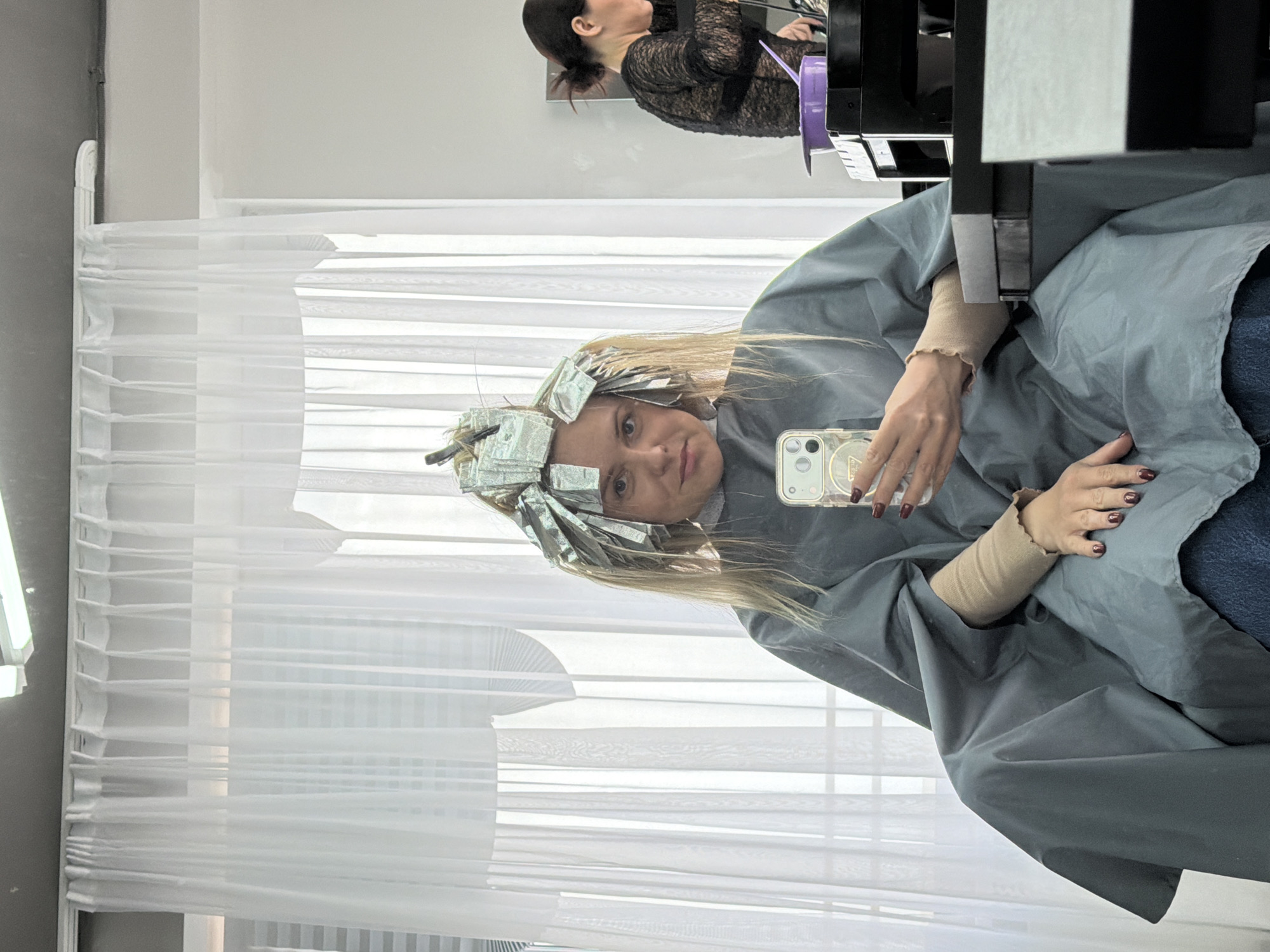 A young woman sits in a salon chair in Votkinsk, Russia, undergoing a hair coloring process. Her hair is covered in foil packets, suggesting highlights or a similar technique. She holds a smartphone in her right hand, taking a selfie or using it for other purposes. She is draped in a light blue or gray salon cape. Another woman, potentially a stylist, stands behind her, slightly out of focus, possibly attending to her hair. In the background, a partially visible window has sheer, white blinds. The overall environment is clean and modern.