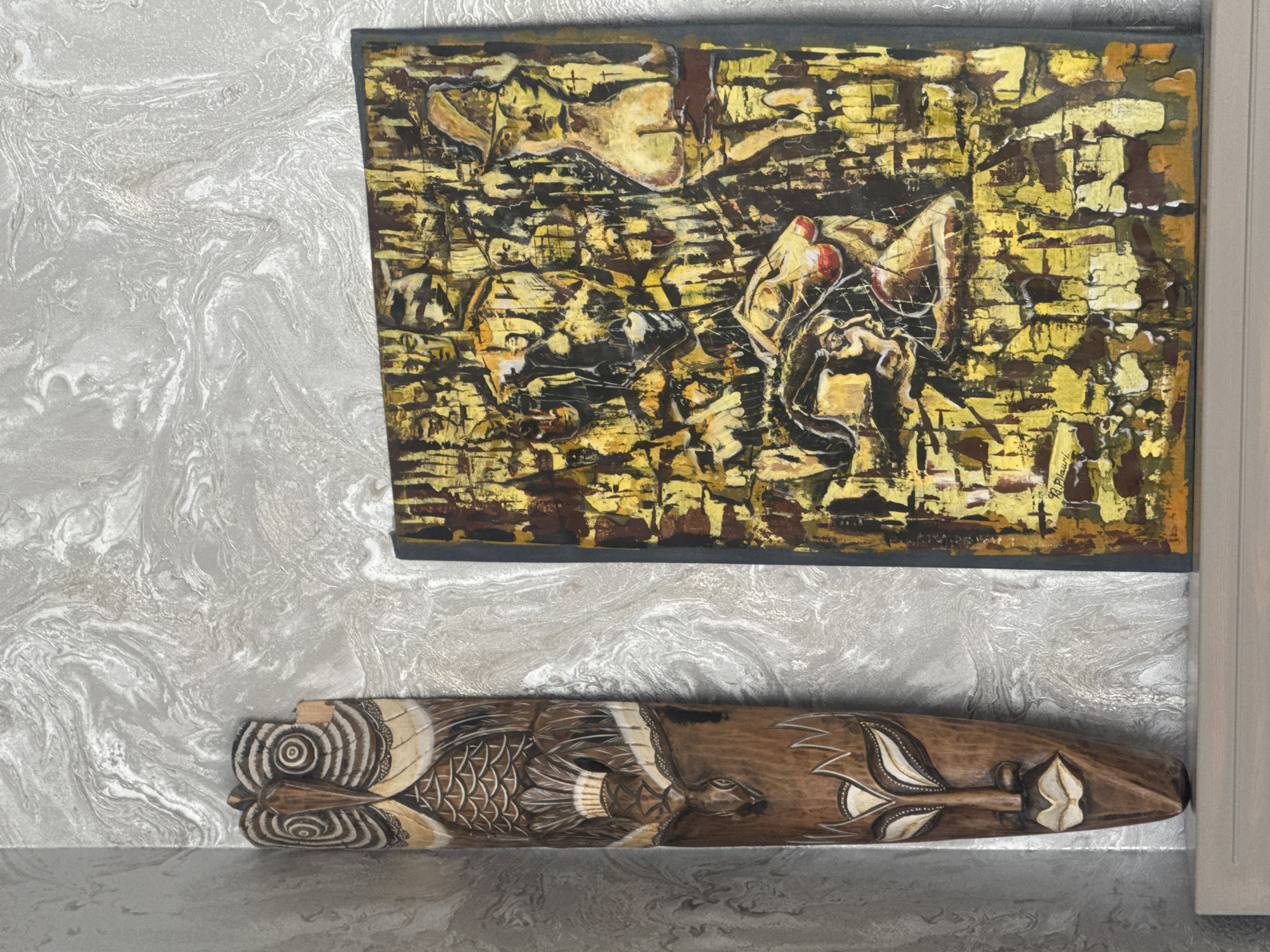An abstract painting and a carved, wooden mask are displayed indoors against a gray marble-patterned wall. The painting, framed in dark gray, features a textured composition of gold, brown, and black hues, with indistinct figures possibly human. Below the painting rests a brown, wooden mask carved with stylized fish and foliage designs, positioned horizontally. The setting appears to be an interior space with a muted gray color scheme, evidenced by a partial view of a doorframe on the right. The location is presumed to be Tbilisi, Georgia.