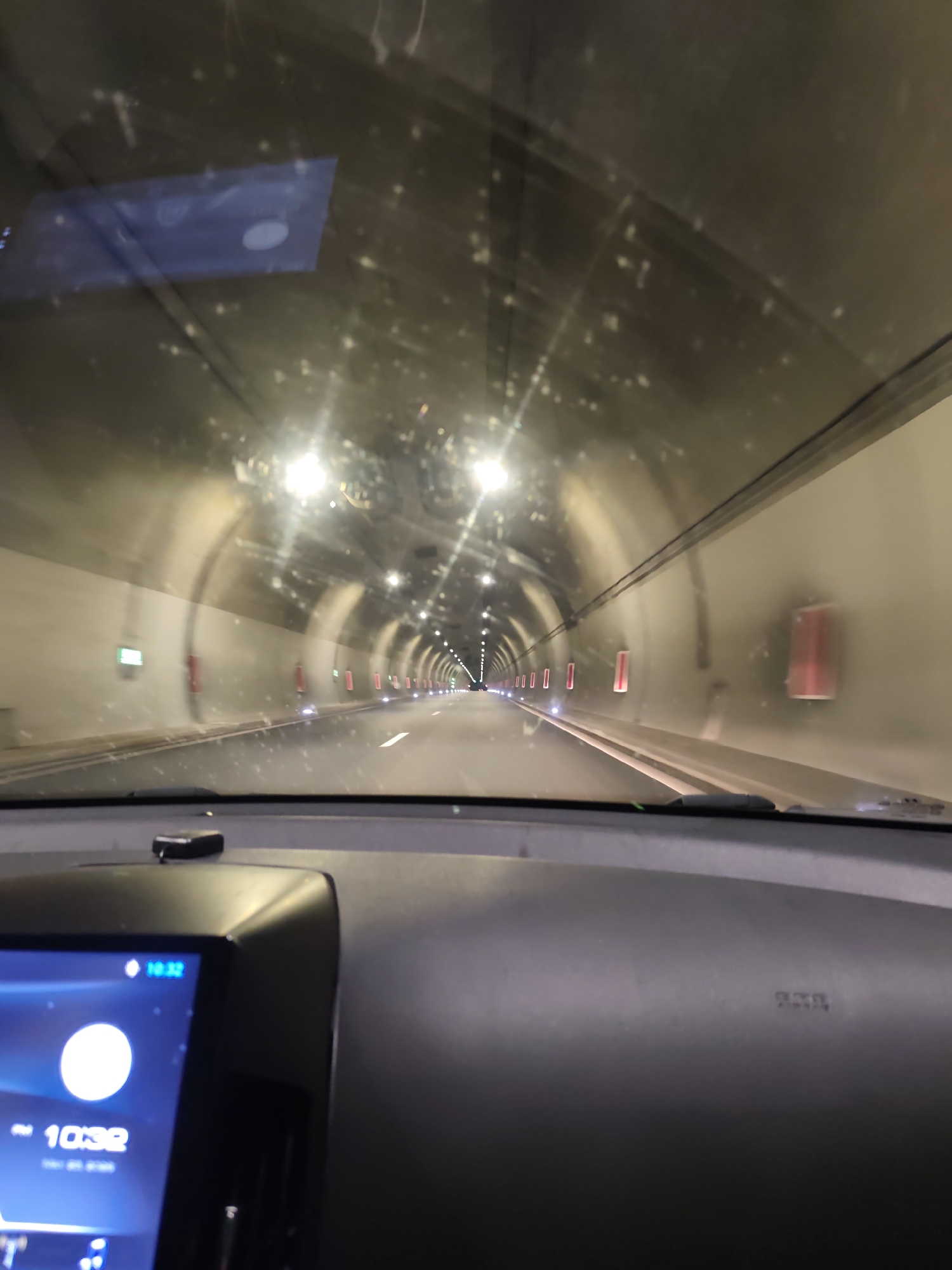 The image shows a car driving through a tunnel. The dashboard is visible, with a digital clock displaying 10:32. The tunnel has lights and emergency exits along the walls. The windshield is slightly dirty. It is the Jeleznitsa tunnel, located in Jeleznitsa, Bulgaria. No people are clearly visible.