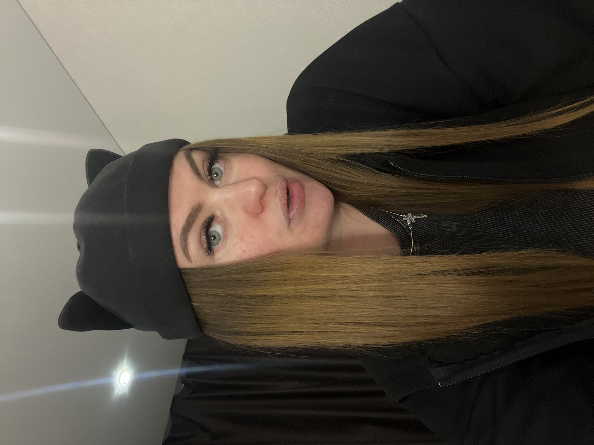 A young woman with light blonde hair is pictured in a close-up shot. She is wearing a black hat with cat ears and a black jacket. Her hair falls across her face, partially obscuring her right eye. She is also wearing a silver necklace. The background consists of a white wall. The location is Unknown, Unknown.