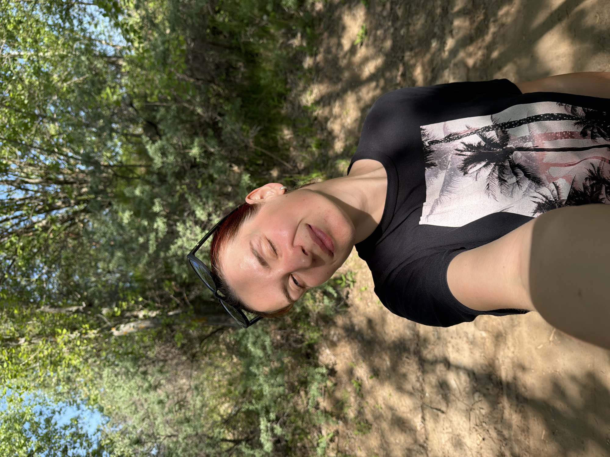 A woman is posing for a selfie. She is outside on a dirt path surrounded by greenery. She is wearing a black t-shirt with a palm tree design and has sunglasses perched on her head.