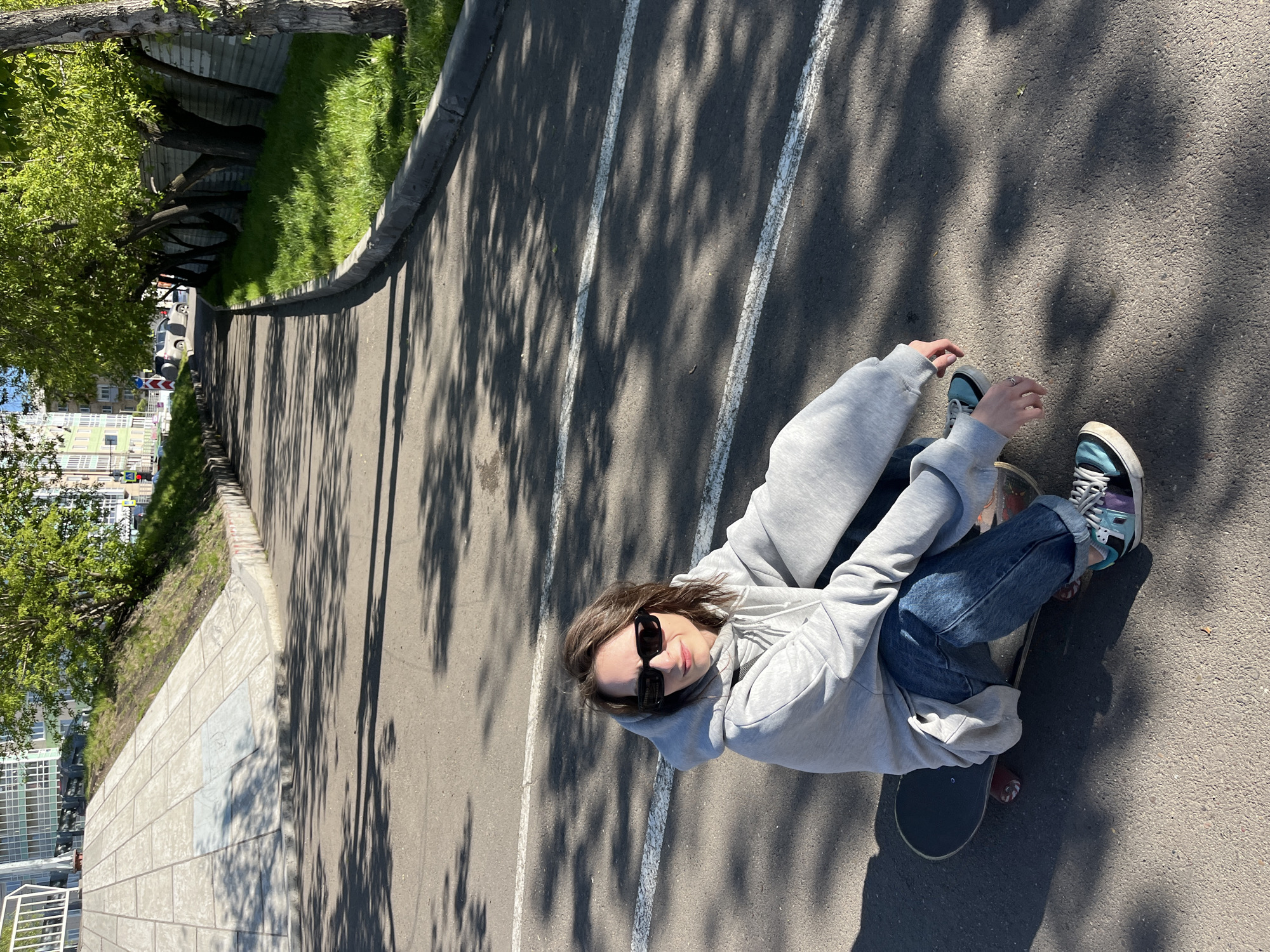 A young person wearing a gray hoodie, blue jeans, and multicolored sneakers is sitting on a skateboard on an asphalt surface with white painted lines and shadows from trees. The person also has sunglasses. The location appears to be an outdoor urban area, likely a city street or pathway with vegetation and buildings visible in the background.