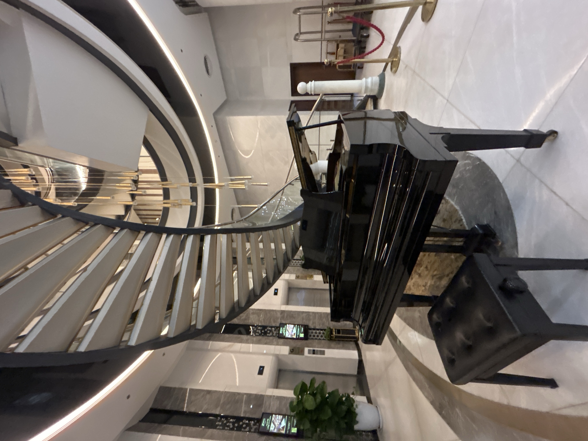 The image depicts an indoor scene featuring a black grand piano with its matching bench, positioned near a curved staircase. The staircase's design includes white balusters and dark, linear accents. Behind the piano, there are elevators and potted plants. The surrounding environment appears to be a modern building, likely a hotel or an upscale commercial space. Based on the image location information, it is likely this scene is in Bac Nha Trang Ward, Vietnam. No individuals are present in the image.