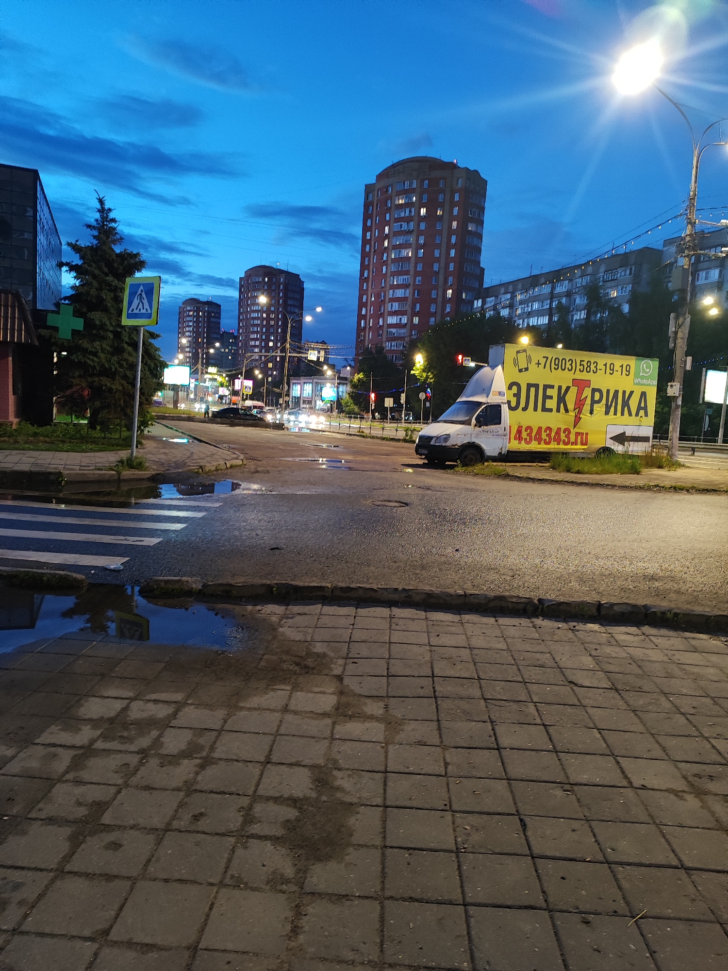 The image shows a street scene at dusk or night in an urban environment. In the foreground, a pedestrian crossing is visible with wet pavement reflecting the blue twilight sky and surrounding lights. To the right of the crossing, a white van with a large yellow advertisement is parked. The advertisement features a lightning bolt symbol and Russian text, "ЭЛЕКТРИКА" (Elektrika) along with a phone number and a website address. Behind the van, tall residential buildings are illuminated with lights from their windows. A street lamp with a bright light is prominently visible in the upper right corner of the image. A green cross symbol, possibly indicating a pharmacy, is also visible on the left side of the image. The sky is a deep blue with some scattered clouds.