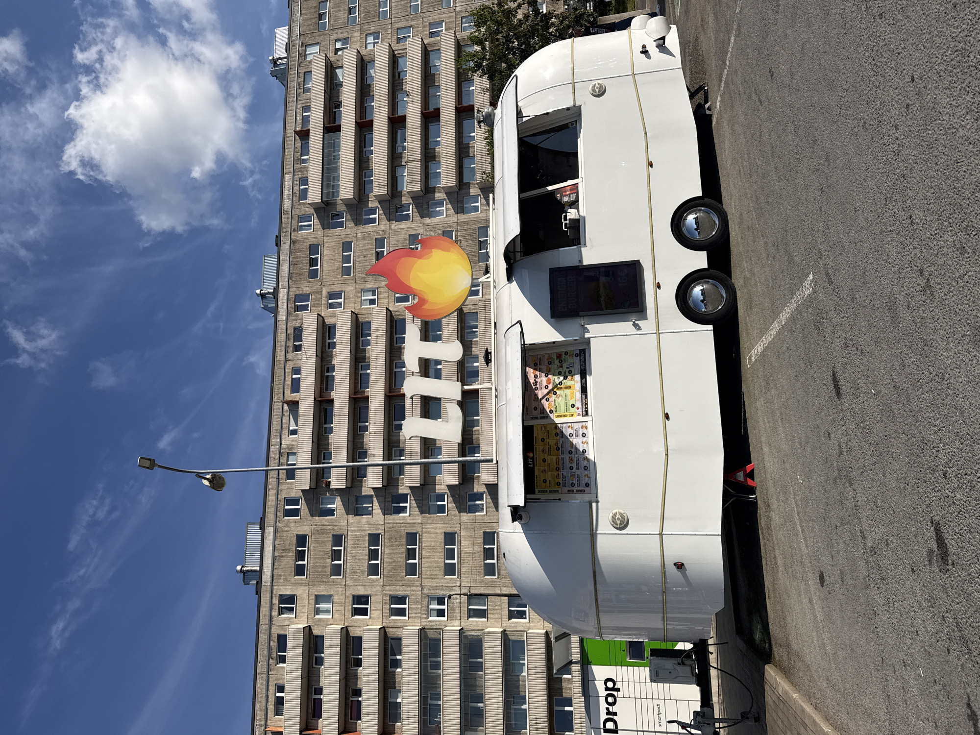 A food truck, labeled "LIT" with a flame logo, is parked in a lot in front of a Soviet-era apartment building. The building is grey concrete with multiple balconies. The food truck is white and rounded, resembling a vintage trailer. It has menus displayed in the windows. To the left of the food truck is a green and white parcel locker labeled "Drop" and "smartpost". The sky is blue with some white clouds. The scene appears to be a street or parking lot in Tallinn, Estonia.