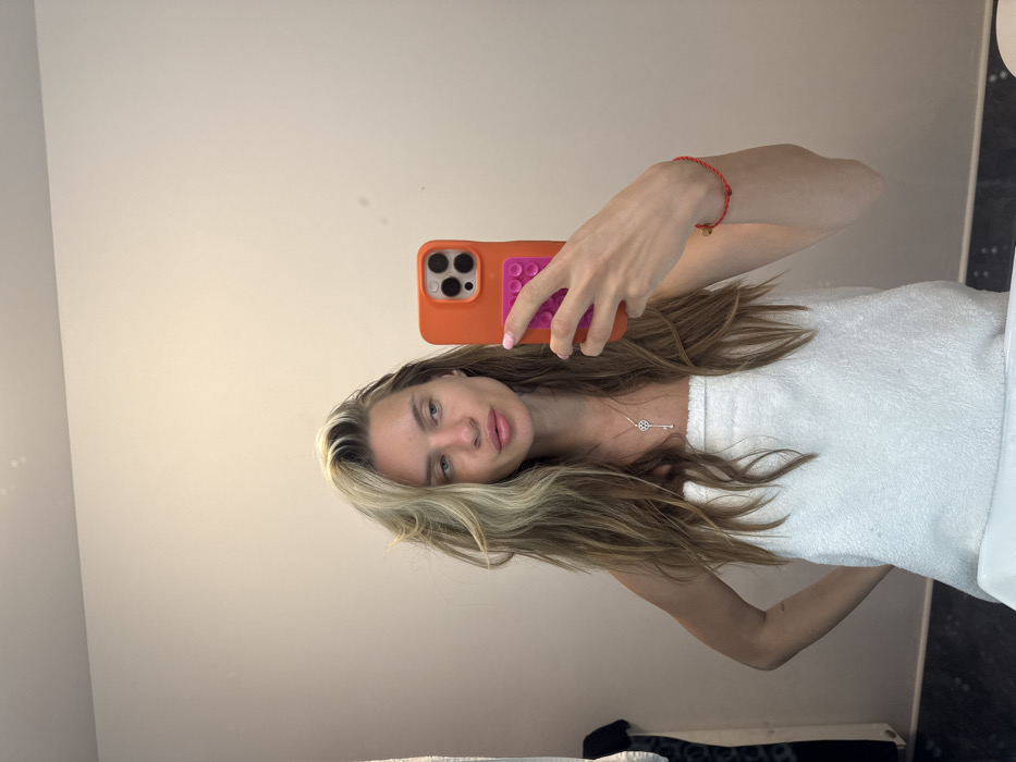 A woman takes a mirror selfie. She has long blonde and brown hair, and is wrapped in a white towel. She wears a delicate silver necklace and a red string bracelet. Her phone has a bright orange case with pink grips. The background is a plain, light-colored wall.