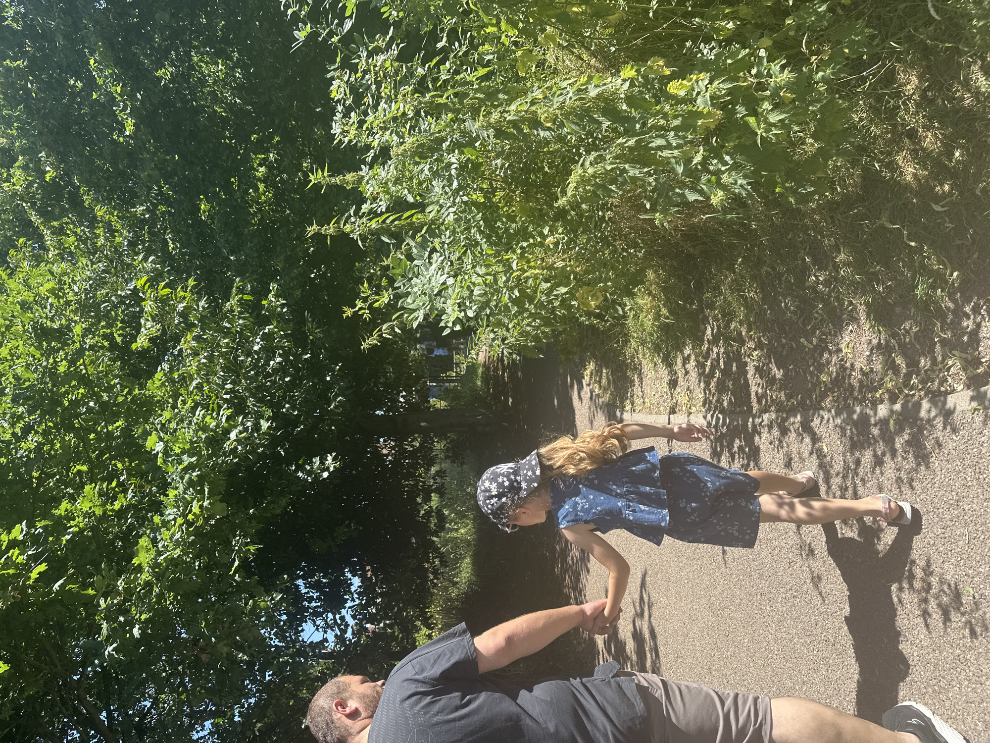 In a park in Trafford, United Kingdom, a man and a young girl are walking hand-in-hand on a paved path. The man, seen from behind, is wearing a black t-shirt and grey shorts. The girl, also viewed from behind, has long blonde hair and is wearing a blue floral dress and a patterned cap. She is wearing pink sandals. Lush green trees and dense vegetation line the path, with dappled sunlight filtering through the leaves. The scene is peaceful and suggests a leisurely stroll on a sunny day.