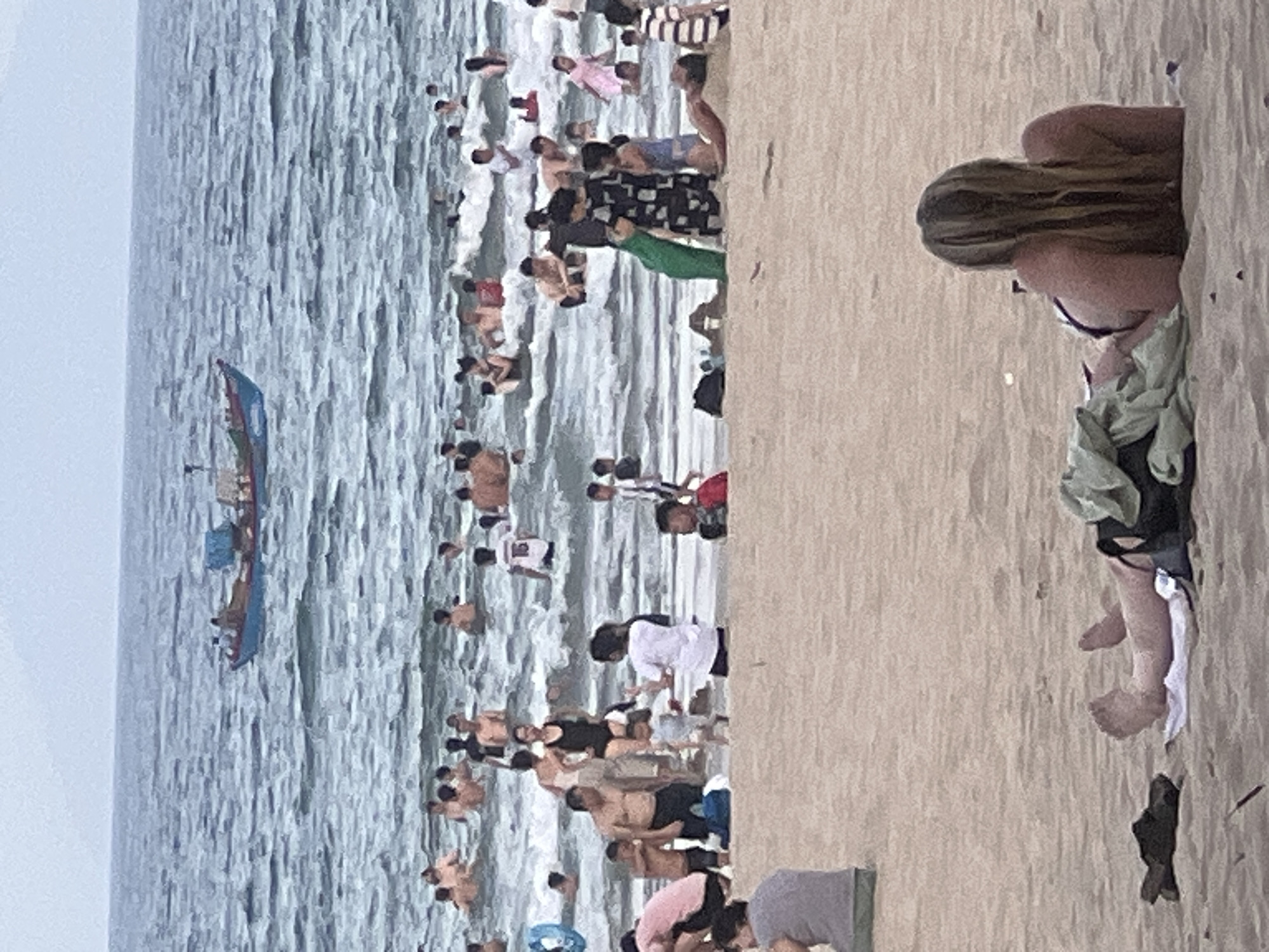 The image depicts a crowded beach scene in Đà Nẵng, Vietnam, likely during the daytime. The ocean occupies the left side of the frame, with visible waves and a blue boat with red and white accents navigating the water. The beach itself is filled with a multitude of people, suggesting a popular recreational area.
Individuals are engaged in various beach activities. Some are in the water, wading or swimming, while others are standing or sitting on the sandy shore. The people are dressed in swimwear, casual clothing, and some are holding or draped with towels or sarongs. The atmosphere appears lively and social, typical of a summer day at a busy beach.
Notable details include the diverse attire of the beachgoers, ranging from patterned swimsuits to simple t-shirts and shorts. The sand appears light in color, and the water is a muted blue-grey, indicating it might be a slightly overcast day or the lighting conditions are affecting the water's hue. The presence of the boat in the water suggests it could be a fishing boat or a small passenger vessel. There is no visible text in the image.