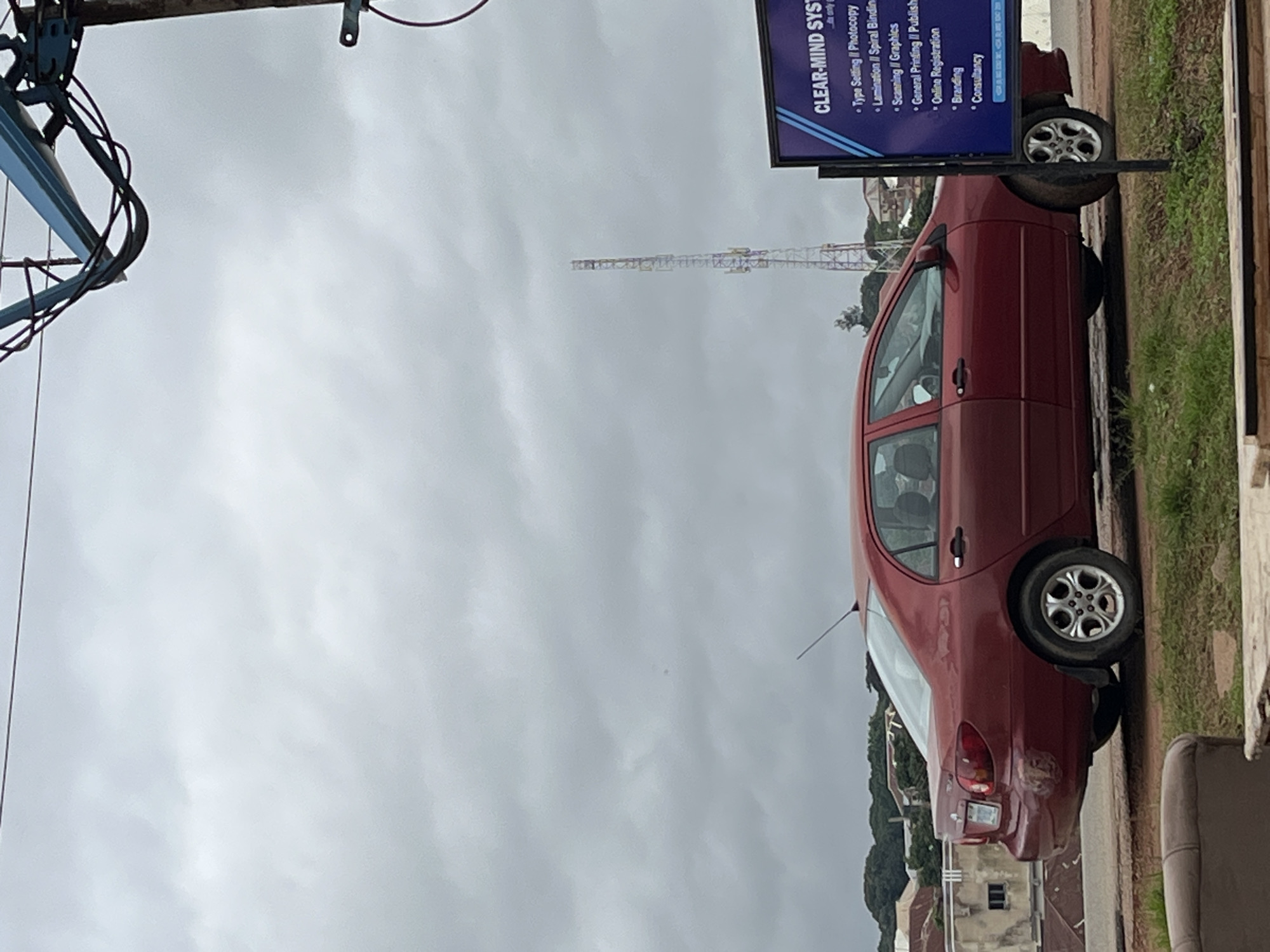 The image shows a red sedan parked on the side of a road in Jos, Nigeria. The car is facing right and appears to be a Toyota Corolla. The sky is overcast with a communication tower visible in the distance. A sign is partially visible on the right side of the image, reading "CLEAR-MIND SYST...". The foreground shows a brown sofa and some grass. The background reveals buildings and trees, characteristic of an urban or semi-urban setting in Jos. The overall scene suggests a cloudy day, possibly before or after rain. No people are visible in the image.