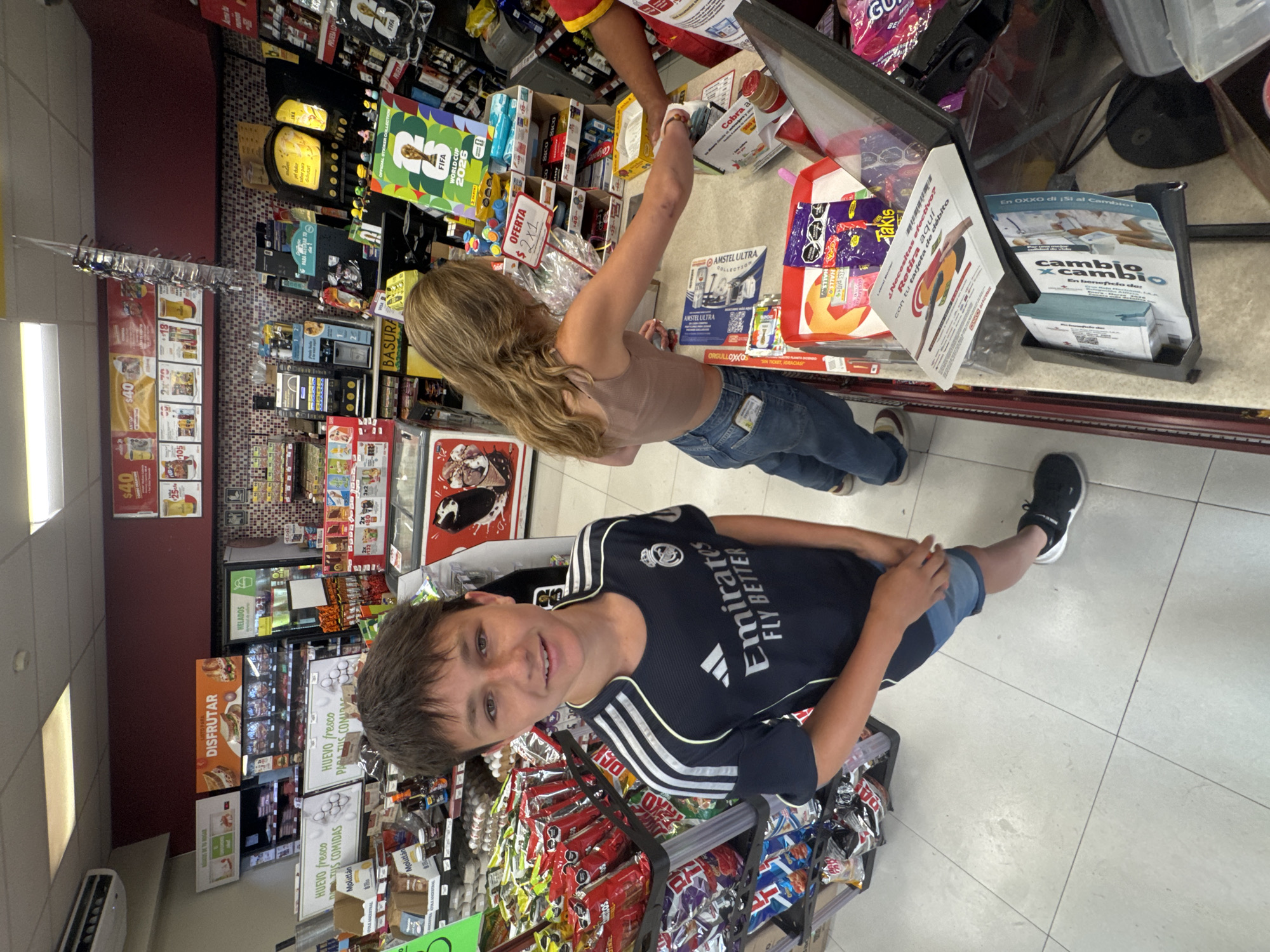 The image depicts a scene inside an OXXO convenience store, likely in San Bernardino Tlaxcalancingo, Mexico. A young boy wearing a Real Madrid jersey is standing in the foreground, holding a shopping cart filled with snacks. Behind him, a young woman with long blonde hair is at the checkout counter, interacting with a store employee. The shelves behind them are stocked with a variety of products, including snacks, drinks, and household items. There is promotional signage on the shelves. Several advertisements are visible near the cashier area. The floor is tiled.