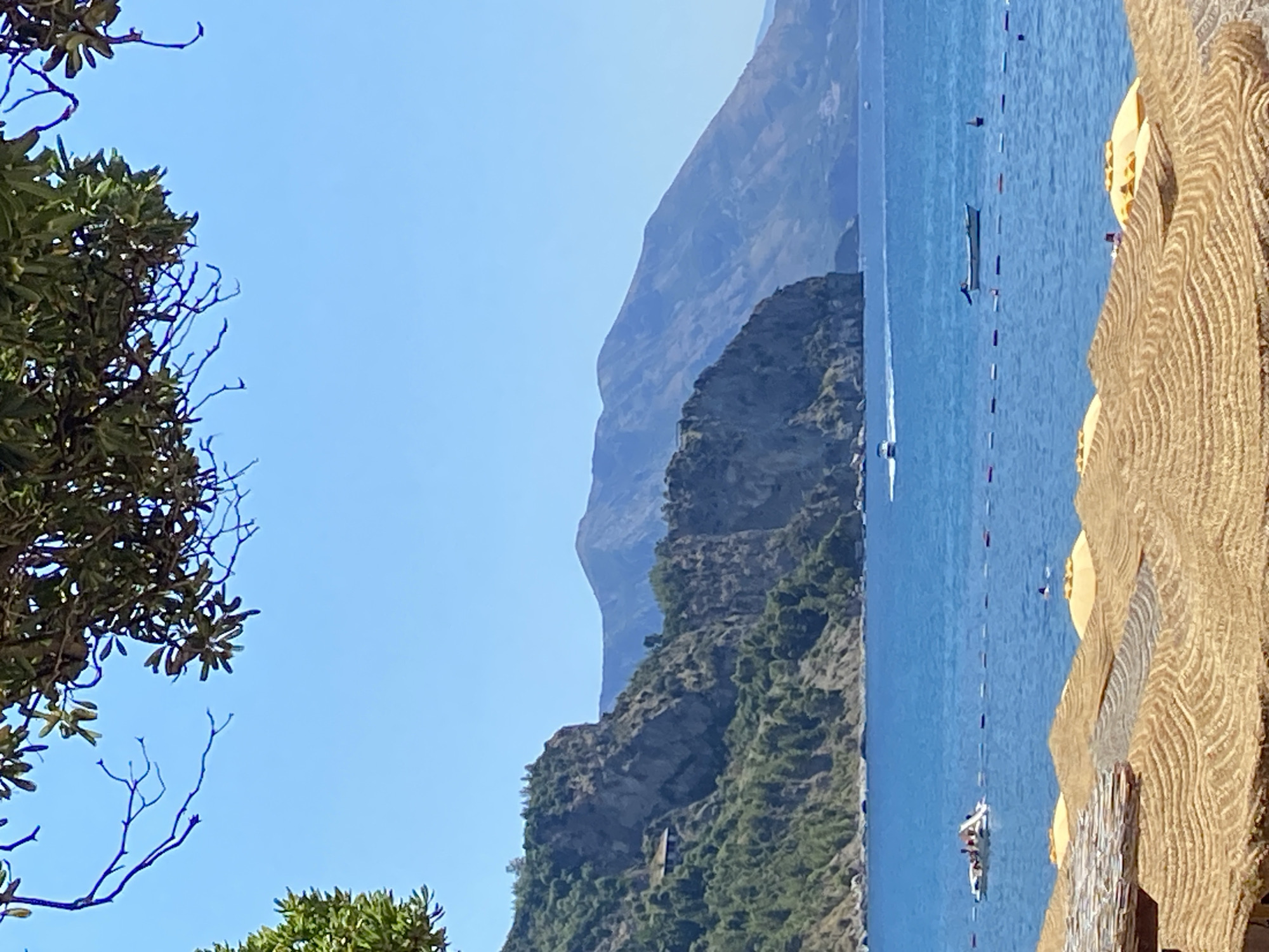 The image shows a coastal landscape with a body of water, rocky hills, and vegetation. In the foreground, there are several thatched umbrellas and what appears to be a wooden structure. The water is a vibrant blue and has several boats on it, including a small white boat with people and a longer, darker boat. A line of buoys stretches across the water. In the mid-ground, a large, rugged cliff face covered in green vegetation rises from the sea. A small building is visible on the side of the cliff. In the background, a mountain range with a hazy, blue hue is visible against a clear blue sky. The scene suggests a warm, sunny day at a beach resort, likely in a Mediterranean or similar climate. This is a view of the coastline near Prijevor, Montenegro.