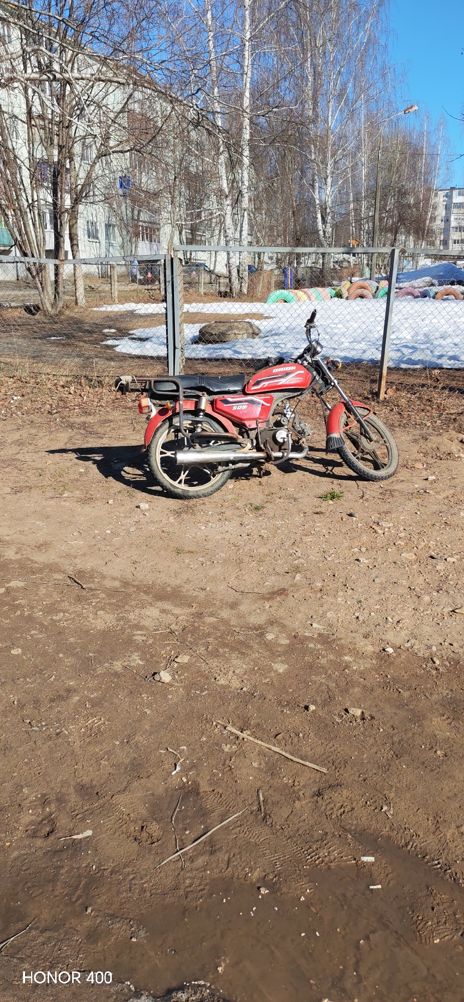 A red Orion 50Q motorcycle is parked on a dirt patch. A chain-link fence runs behind the motorcycle, separating the dirt patch from a snow-covered area with tires used as playground equipment. Buildings are visible in the background, partially obscured by leafless trees. The lower portion of the image shows wet, muddy ground. The word "HONOR 400" is written in the lower left-hand corner. The scene appears to be in Noviy, Russia.