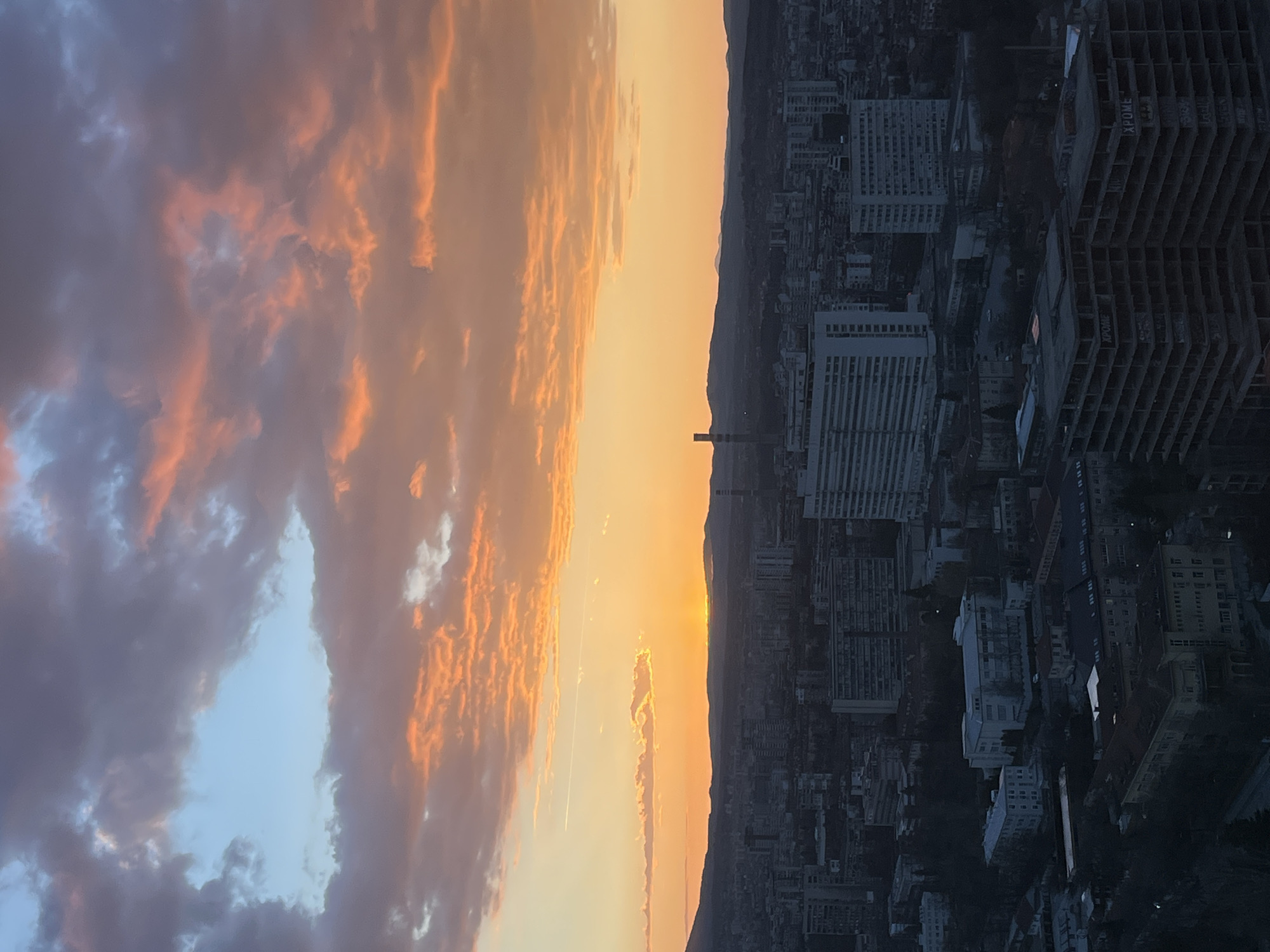 The image presents a view of Sofia, Bulgaria, at sunset. The sky is predominantly orange, with darker, cloudier areas transitioning to a blue tint on the left side. The city is on the right side of the image, with numerous buildings of various heights. A single tall smokestack stands out against the horizon where the sun is setting. The cityscape is darker, suggesting a lower light level compared to the bright sky.