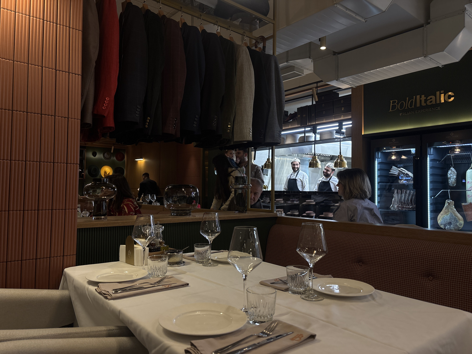 This media file presents the inviting interior of an upscale Italian restaurant, "Bold Italic," located in Yerevan, Armenia. The scene captures an ambient evening setting, characterized by warm, dim lighting.
In the foreground, a dining table is meticulously set with a crisp white tablecloth, white plates, polished silverware, wine glasses, and water glasses, awaiting guests. A beige upholstered dining chair is partially visible on the left. The restaurant's decor includes a striking vertical wall paneling of reddish-brown ridged tiles on the far left. A unique design element is a rack of men's suit jackets in various colors, hanging above the dining area, adding a sophisticated, sartorial flair.
Through a large glass partition in the background, a lively kitchen is visible, where several chefs in white uniforms and hats are at work, some seen smiling and interacting, indicating a bustling environment. To the right of the kitchen, large refrigerated display cases, clearly labeled "DRY AGER," showcase an array of premium cuts of meat, including dry-aged beef and what appears to be cured ham or prosciutto, emphasizing the restaurant's focus on high-quality ingredients. A woman with short hair is seated at a booth to the right, facing towards the kitchen and dry-aging units.
The restaurant's branding, "Bold Italic," is prominently displayed in elegant gold lettering on a dark green textured wall, accompanied by the tagline "ITALIAN EXPERIENCE." Other visible details include industrial-style ceiling ventilation ducts, pendant lights illuminating the kitchen pass, and a subtle "B" or "BI" logo on the napkins. The overall atmosphere conveys a refined and welcoming dining experience centered around Italian cuisine and quality meats.