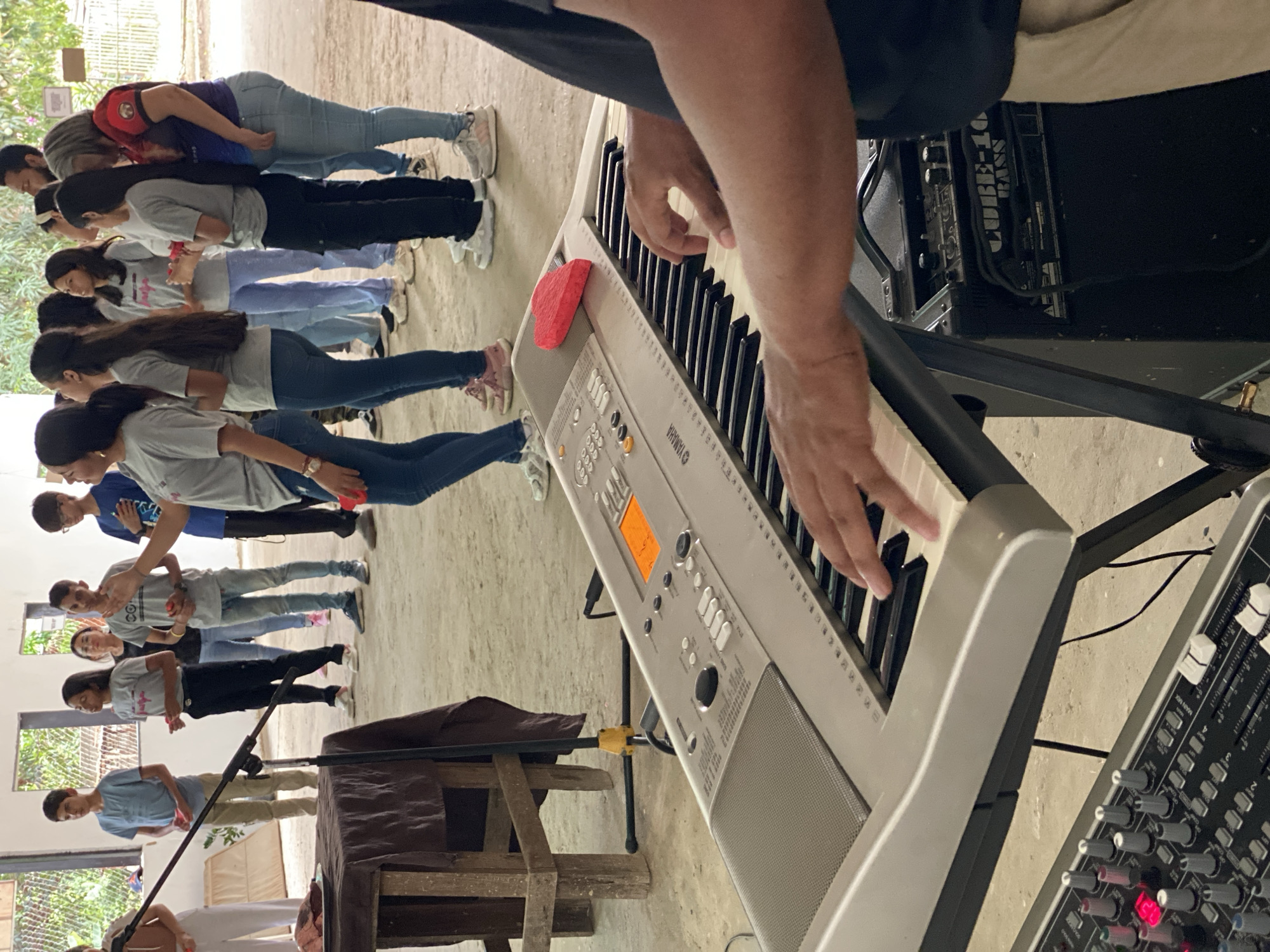 The image shows a group of people lined up in what appears to be an indoor space, likely a community hall or school in Tinaquillo, Venezuela. A keyboard player is in the foreground, focused on playing the keyboard. The people are lined up with their heads bowed. The environment looks simple, with a cement floor. There is some basic equipment present such as an amplifier and sound board. There are some windows in the background, as well as a table.