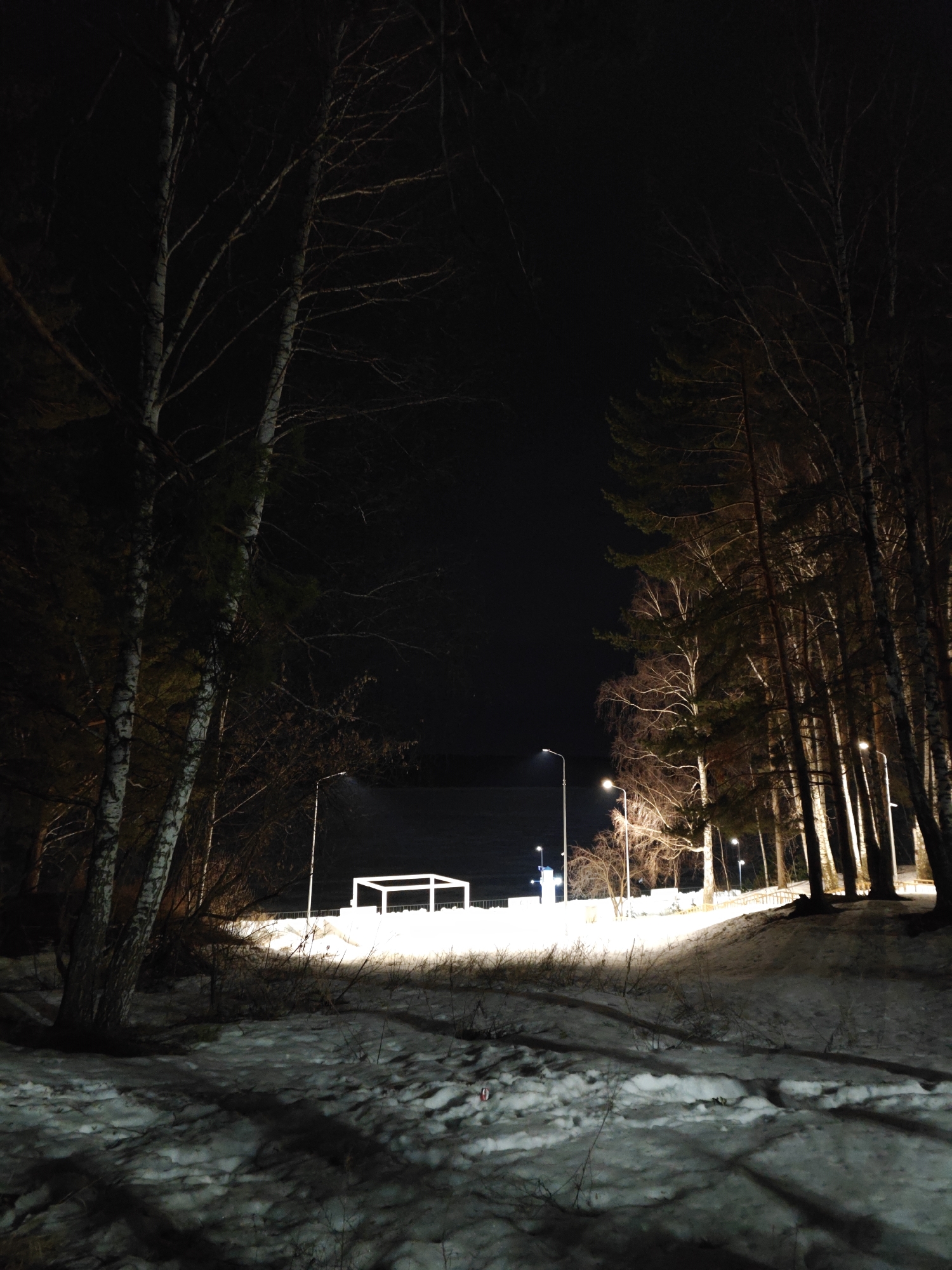 The image depicts a snowy, night scene near the Kama River in Chaikovsky, Russia. Bare trees frame a view toward a brightly lit area on the waterfront. A simple, white cube structure stands out. Several streetlights illuminate a pathway alongside a body of water, the Kama River. The foreground is covered in snow, with scattered shadows from trees and other vegetation.