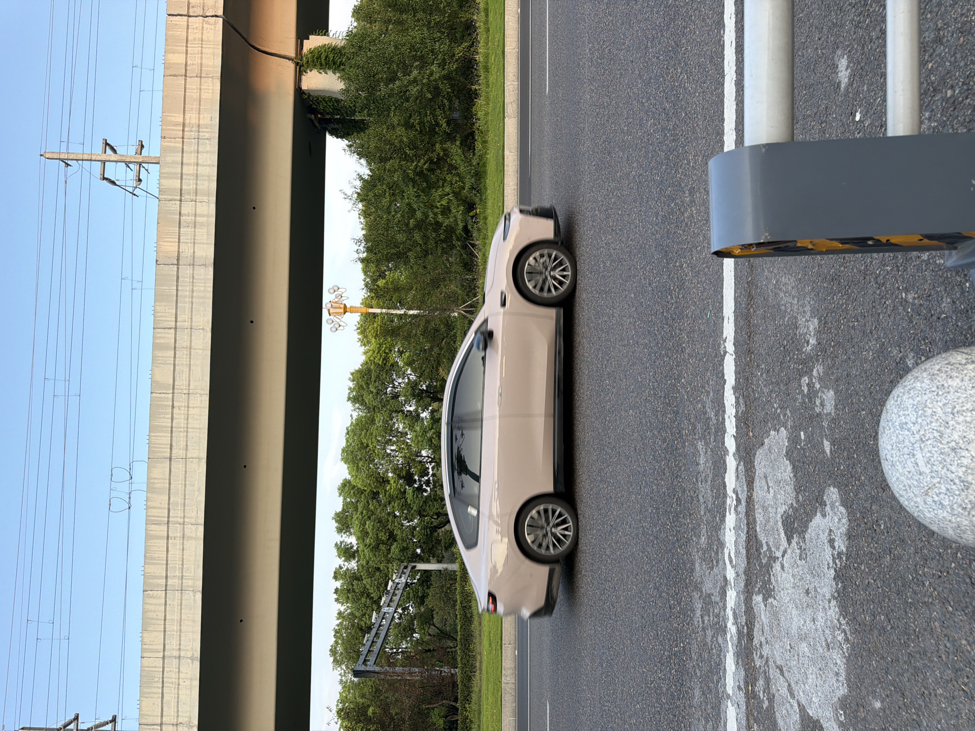 A light-colored sedan is captured in motion on a paved road, likely in Jiangjin, China, given the infrastructure visible. The car is positioned in the center-right of the frame, with its left side facing the camera. Above the road, a substantial concrete overpass is prominent, supported by columns. Overhead, a clear blue sky is crisscrossed with power lines and utility poles. To the left and right of the overpass, lush green trees form a dense backdrop. In the foreground, the road surface shows some wear and tear, with a white lane marking and what appears to be a concrete bollard on the lower left. A metal guardrail with a gray protective covering is visible on the lower right. The overall scene suggests an urban or suburban setting with significant transportation infrastructure.