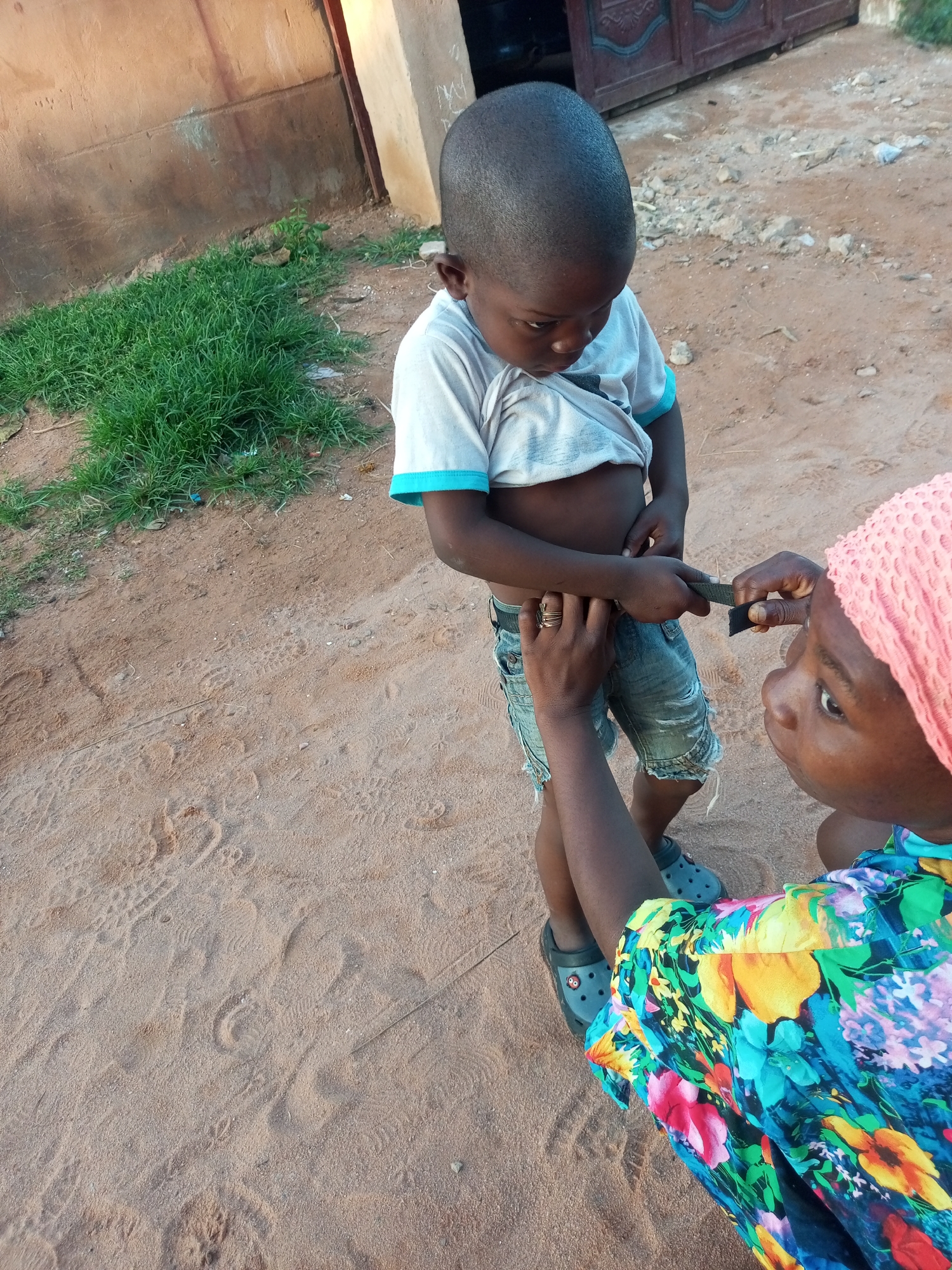 A woman, wearing a pink headscarf and a brightly colored floral shirt, is kneeling on sandy ground. She is attending to a young boy who stands in front of her, wearing a white t-shirt with blue trim and denim shorts. The woman's hands are near the boy's waist, and she appears to be adjusting his belt or shorts. The boy is looking down at his midsection, seemingly focused on the task.
The setting is outdoors, with a sandy, dirt-like ground scattered with pebbles and some dry leaves. To the left of the frame, a patch of vibrant green grass grows beside a weathered, reddish-brown wall. In the background, a dark, ornate metal gate is visible. The lighting suggests it is daytime, possibly late afternoon, given the soft light and shadows. The overall impression is one of a casual, everyday moment in a possibly rural or semi-urban environment. There is no visible text in the image.