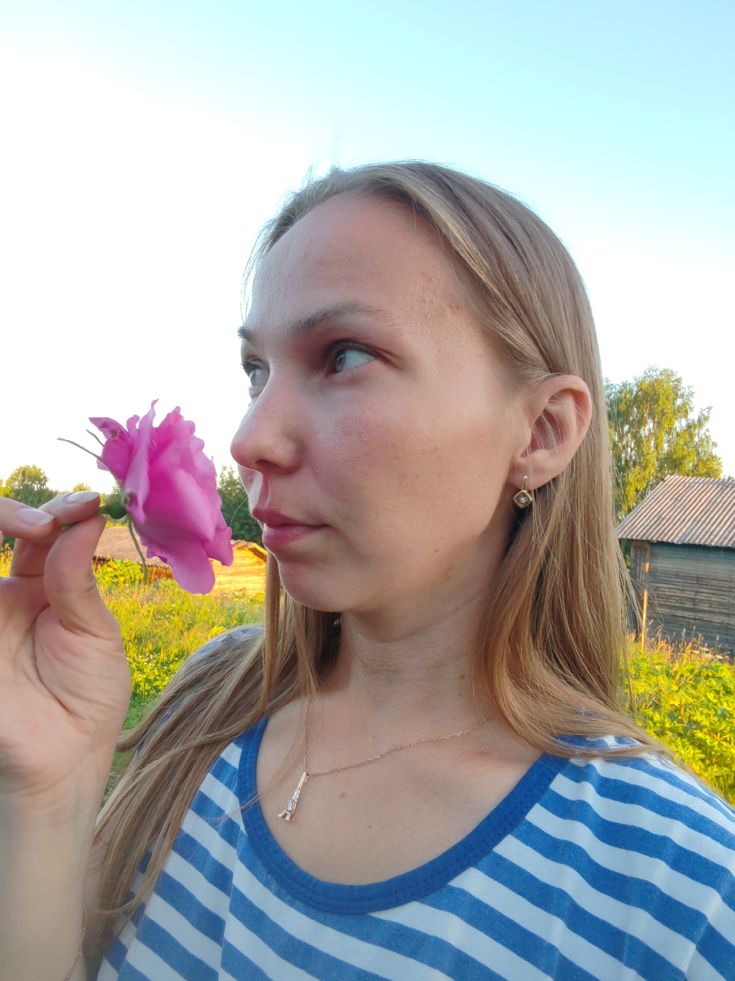 A young woman with long, blonde hair is holding a pink rose up to her nose and smelling it. She is wearing a blue and white striped t-shirt and a delicate necklace with a tower pendant. In the background, there is a wooden building with a corrugated roof and a field of tall grass bathed in sunlight. The sky is clear and blue. The scene appears to be outdoors in a rural setting, possibly in Chuhloma, Russia, given the characteristic wooden architecture and natural landscape.