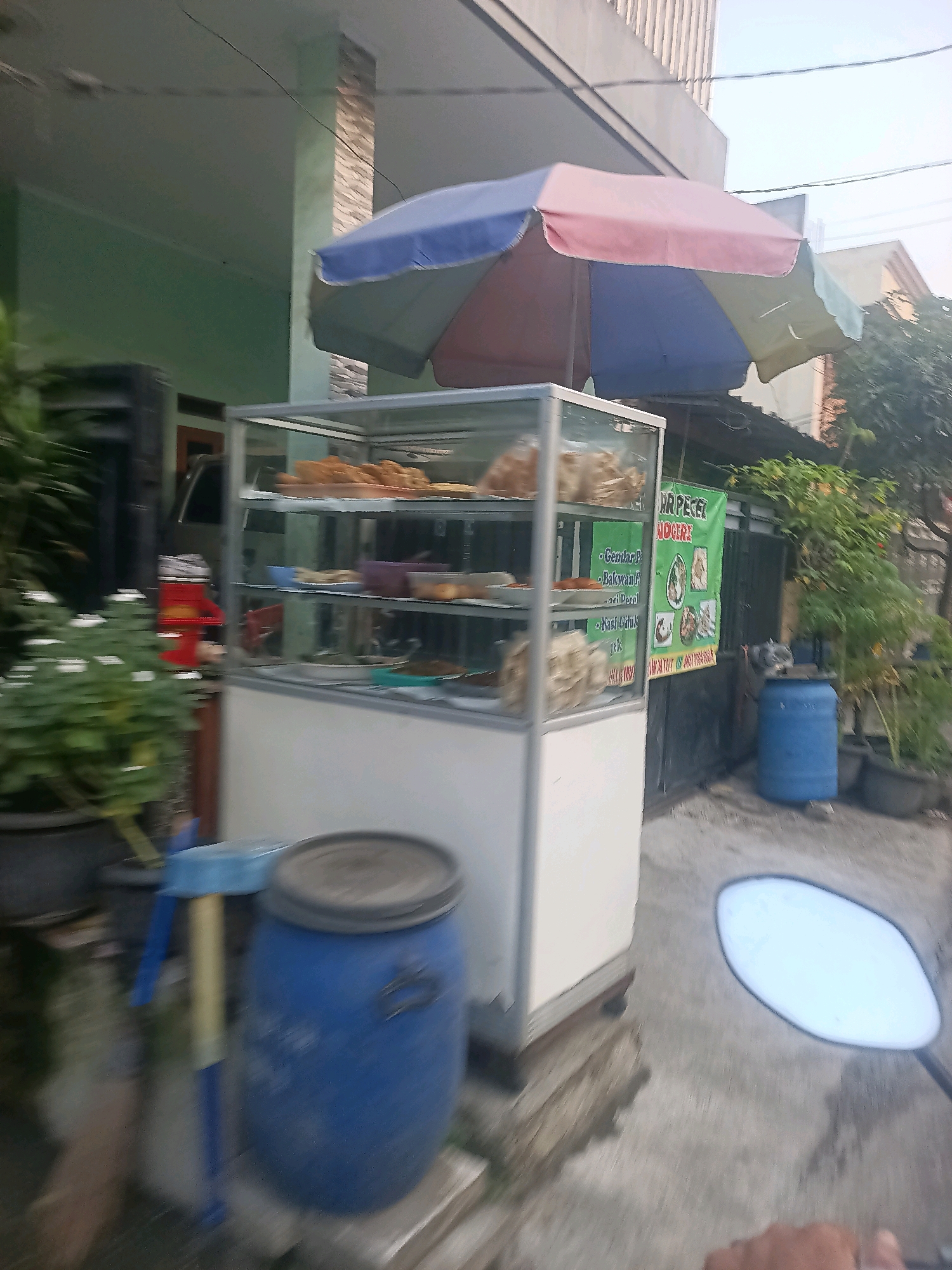 The image depicts a street food stall in Wanajaya, Indonesia. The stall features a glass display case filled with various food items, likely snacks or cooked dishes. Above the stall, a colorful umbrella, with segments of blue, pink, and light yellow, provides shade. To the left of the stall, a potted plant with white flowers is visible, and a red dispenser or container sits behind it. In the background, a green banner with text in Indonesian, including "MR PECEL" and "NOGIRI", is displayed, indicating the name of the food stall or its specialty. Further to the right, a blue plastic barrel and other plants are present. The ground is paved, and what appears to be a motorcycle's side mirror and a hand are partially visible in the lower right corner, suggesting the photo was taken from a moving vehicle. The scene is set outdoors, with buildings and trees forming the backdrop.
