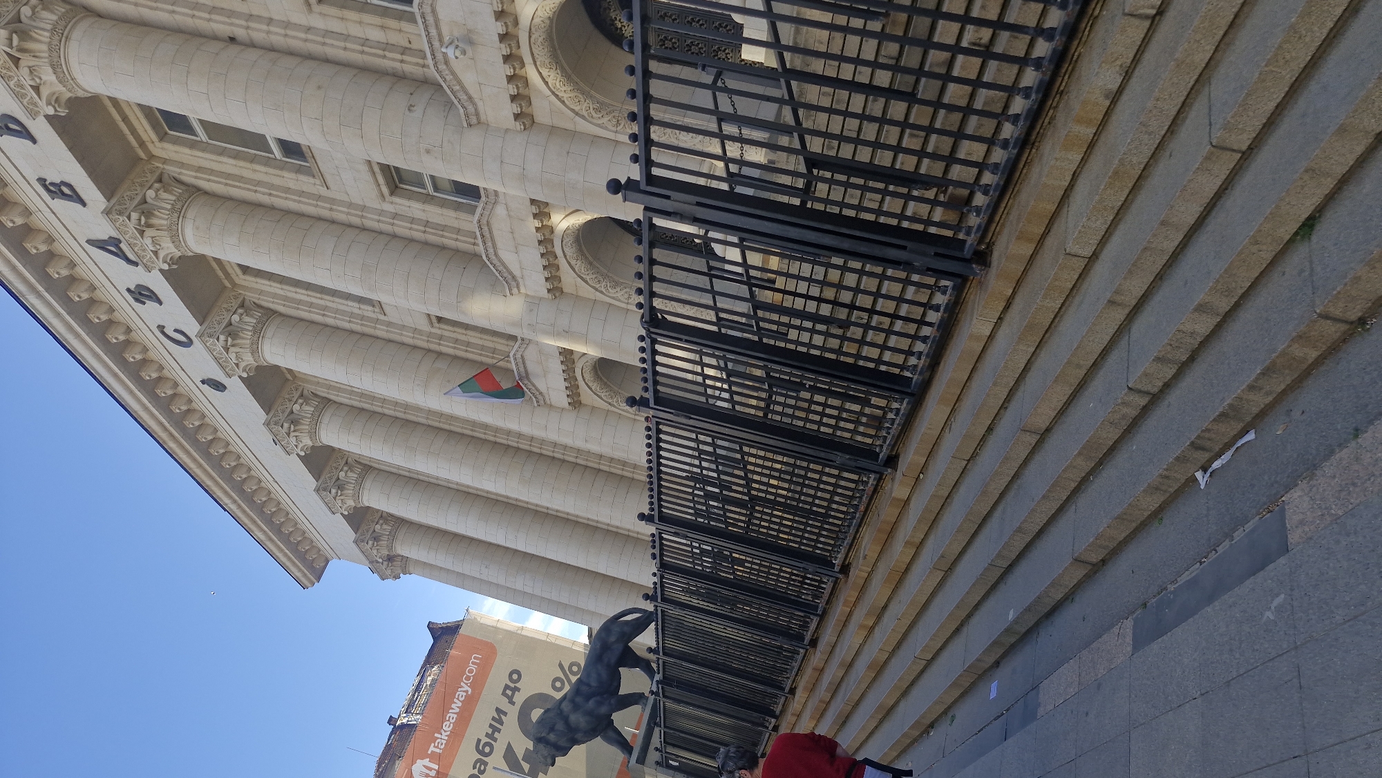 The image shows a view of the National Assembly of Bulgaria in Sofia, Bulgaria. The architecture features classical columns and a balcony with black iron railings. A flag of Bulgaria is visible hanging from one of the windows. Below the building, a bronze statue of a lion stands near the entrance. A sign with "Takeaway.com" is visible on a nearby building. People are also present, some wearing red clothing. The sky is blue and clear. The foreground shows stone steps leading up to the building.