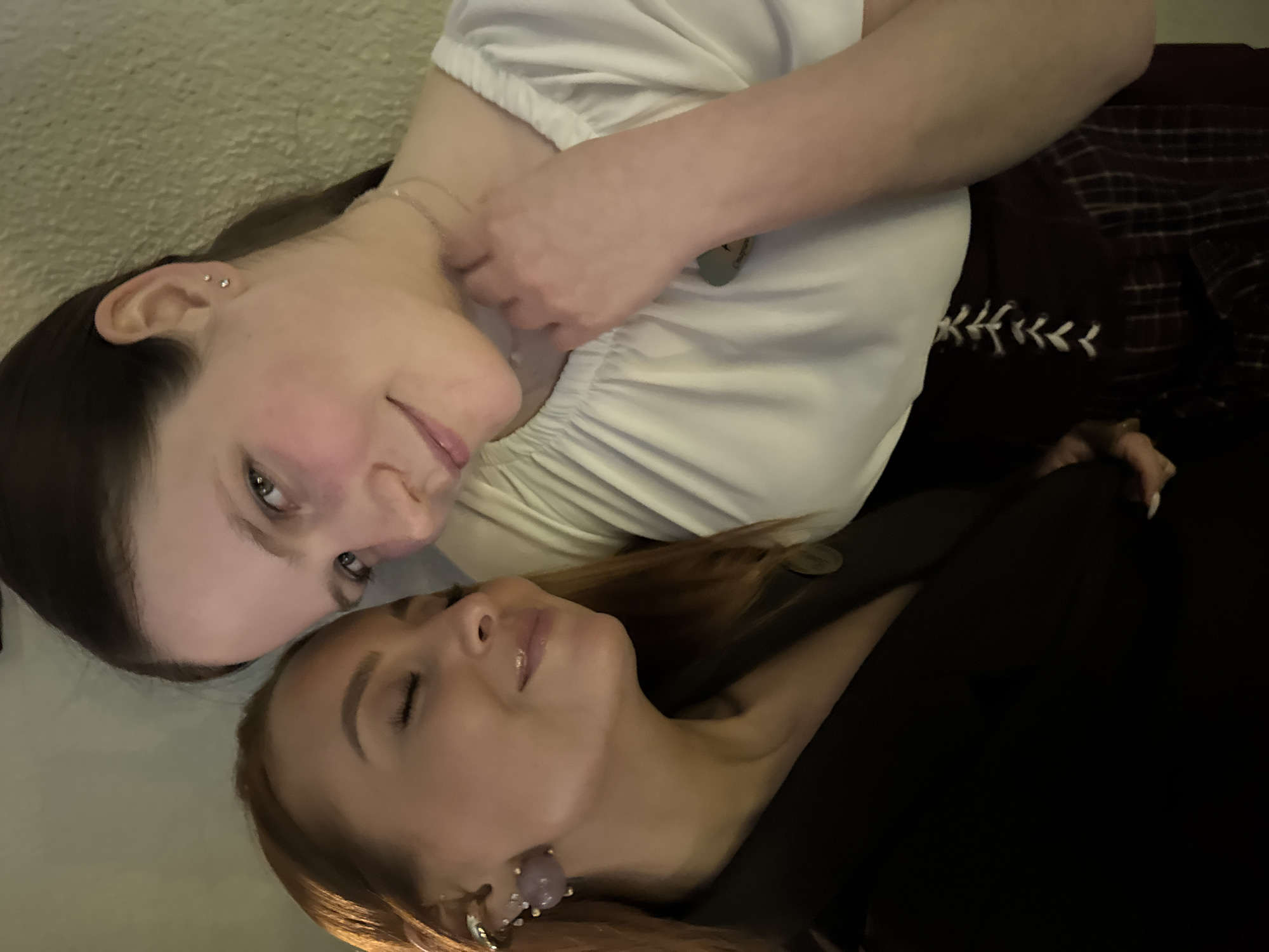 This media file is a portrait-style photograph of two women, taken from a slightly overhead perspective, with the image rotated 90 degrees counter-clockwise from a typical upright orientation. The women are lying down next to each other, their heads close.
The woman whose head is positioned towards the left side of the rotated frame has dark hair, fair skin, and light-colored eyes. She is wearing a white, short-sleeved top with gathered fabric. She has two small silver stud earrings in her visible ear and a delicate chain necklace around her neck. She is looking towards the viewer with a gentle, soft smile. A hand is visible near her chest, and under her arm, a dark garment with a plaid pattern and white embroidered or laced details can be seen.
The second woman, whose head is positioned towards the right side of the rotated frame, has lighter, possibly reddish-brown hair and fair skin. Her eyes are closed, and her expression is peaceful and serene, suggesting rest or relaxation. She is wearing a dark-colored top or dress. A distinct earring, featuring a large purple, possibly stone, component with silver accents, is visible on her right earlobe.
The background behind the first woman is a light-colored, textured surface, possibly carpet or upholstery. The area around the second woman is darker, blending with her dark clothing. The lighting appears to be soft and artificial, indicative of an indoor setting, and creates a sense of intimacy. The overall activity depicted is two individuals resting or relaxing together. There is no visible text in the image. While the location context is Chaikovsky, Russia, there are no specific visual cues within the image itself to confirm this.