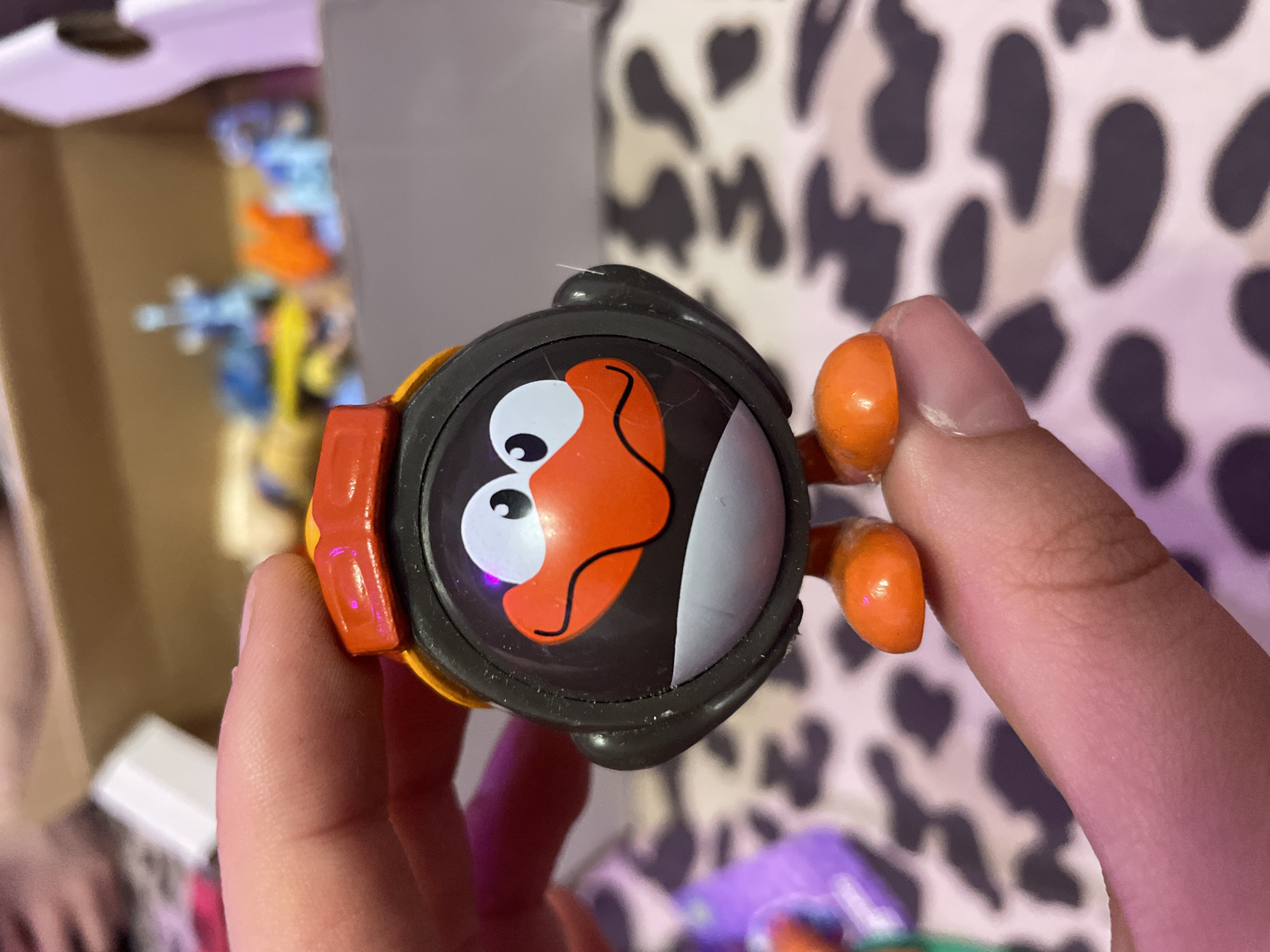 A person's fingers hold a small, black and orange toy penguin with a red helmet. The penguin has a circular body with a cartoonish face featuring large white eyes with black pupils, an orange beak, and a gray chin. The penguin has orange feet and is standing upright. The background is blurry, showing a box with other toys inside, and a surface with a black and white cow-print pattern. There is no information available about the location being Ladozhskii Trudposelok, Russia.