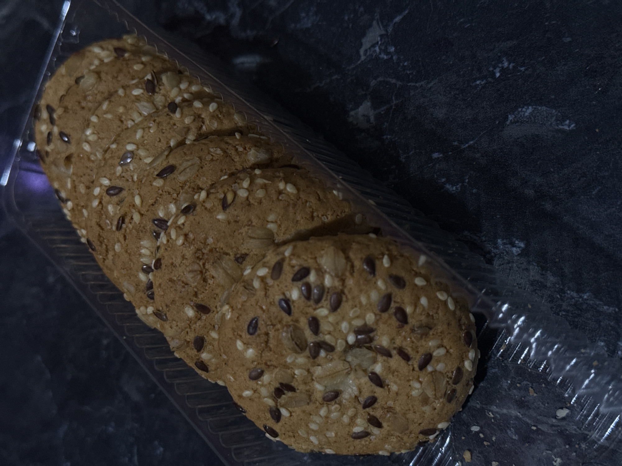 A plastic container holding a stack of round, seeded cookies rests on a dark, marbled surface. The cookies appear to be made of a light brown dough, with visible seeds such as sesame and flax embedded on their surfaces. The container is partially transparent and shows the ribbed texture of its base. The background is dark and indistinct, suggesting an indoor setting. The image was taken in the city of Lobacheve, Ukraine.