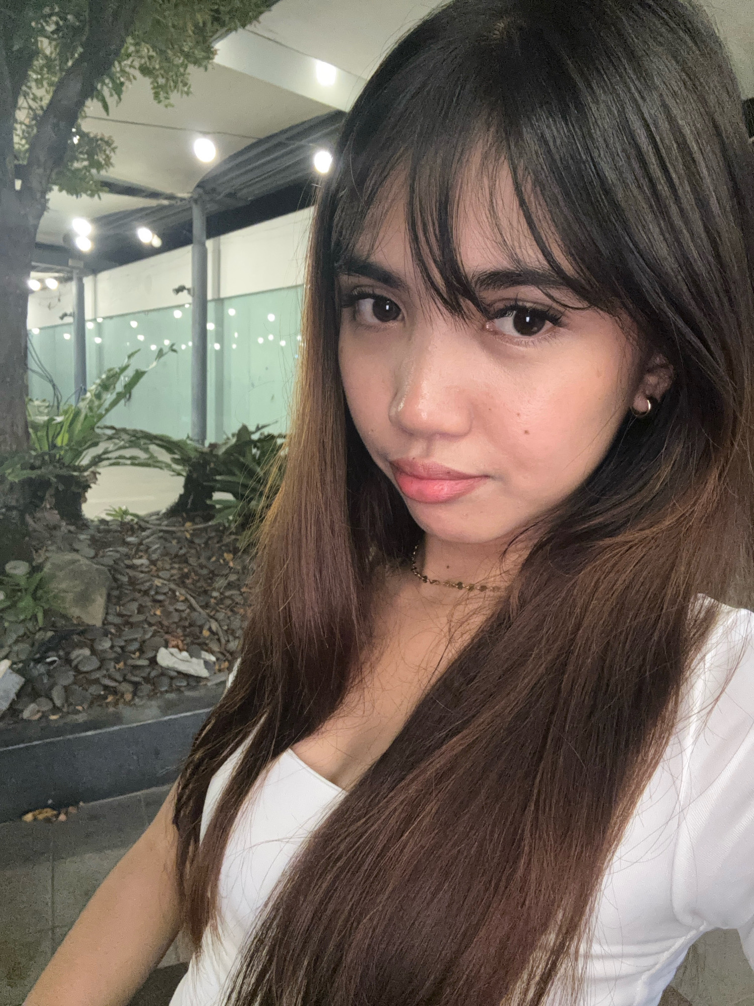 The image is a selfie taken by a young woman with long, dark brown hair and bangs. She is wearing a white top and a gold necklace. She has a small gold hoop earring in her left ear. Her skin is fair with a few freckles. She is looking towards the camera with a slight smile. In the background, there is a tree with green leaves, a patch of ornamental plants, and a gravel-covered ground. String lights are visible in the distance, suggesting an outdoor setting, possibly a restaurant or a garden area. The lighting suggests it is evening or nighttime. The presence of string lights and ornamental plants indicates a possibly urban or semi-urban setting. While the image does not explicitly show any landmarks unique to Davao City, such a scene with ornamental plants and string lights could be found in various establishments or public spaces within the city, such as parks, cafes, or garden restaurants.